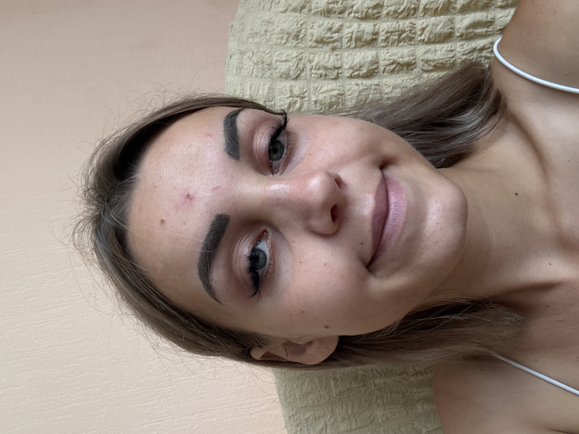 A young woman is pictured from the chest up, looking towards the camera. She has fair skin with a few blemishes on her forehead and cheek. Her eyebrows are dark and well-defined, and she is wearing false eyelashes. Her eyes are open and blue-green. She has a small mole on her right cheek. Her lips are slightly parted in a faint smile, and she is wearing light pink lipstick. Her hair is light brown and falls to her shoulders. She is wearing a white camisole with thin straps. The background is a textured, beige wall and a yellow patterned couch or chair. The lighting is soft and natural, suggesting it was taken indoors during the day. There is no indication of the city of Kholmsk, Russia, or any other specific location within the image.