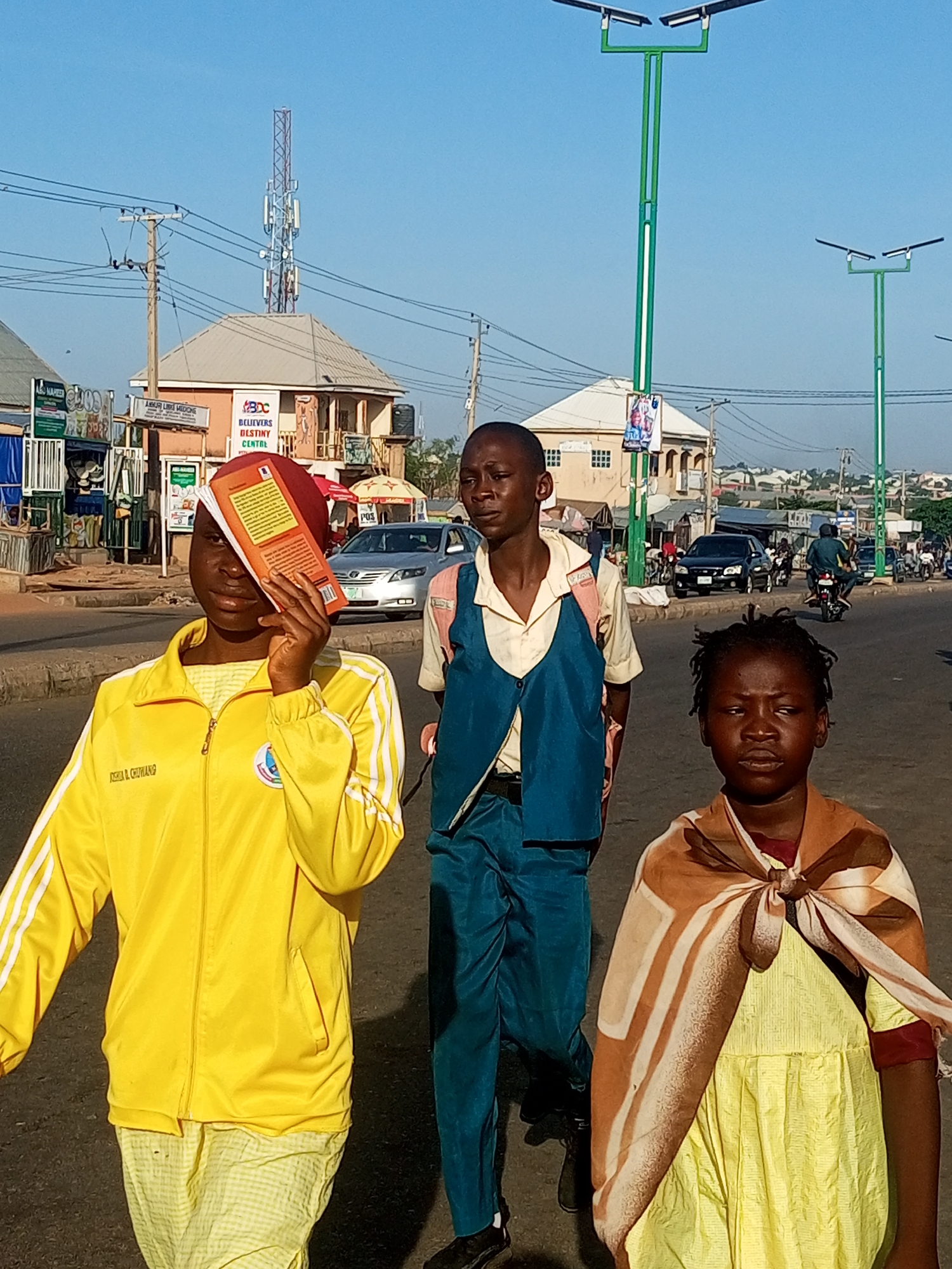 A medium shot captures three young individuals walking on a paved street under a clear blue sky, suggesting a daytime scene in Tempure, Nigeria. The individual on the left, a young woman, wears a bright yellow tracksuit with white stripes and holds an orange book in front of her face, obscuring it from view. She also wears a patterned yellow garment underneath the tracksuit. Next to her, a young man dressed in a light-colored shirt, a dark blue vest, and dark blue trousers walks forward, carrying a light-colored backpack. On the right, a young person wears a yellow dress adorned with a patterned brown and white scarf tied around their neck and shoulders. In the background, buildings with red and brown facades line the street, interspersed with shops and advertising signs. Traffic, including cars and motorcycles, moves along the road, and overhead power lines crisscross the sky. The environment appears to be an urban or semi-urban area, characteristic of Nigerian street scenes.