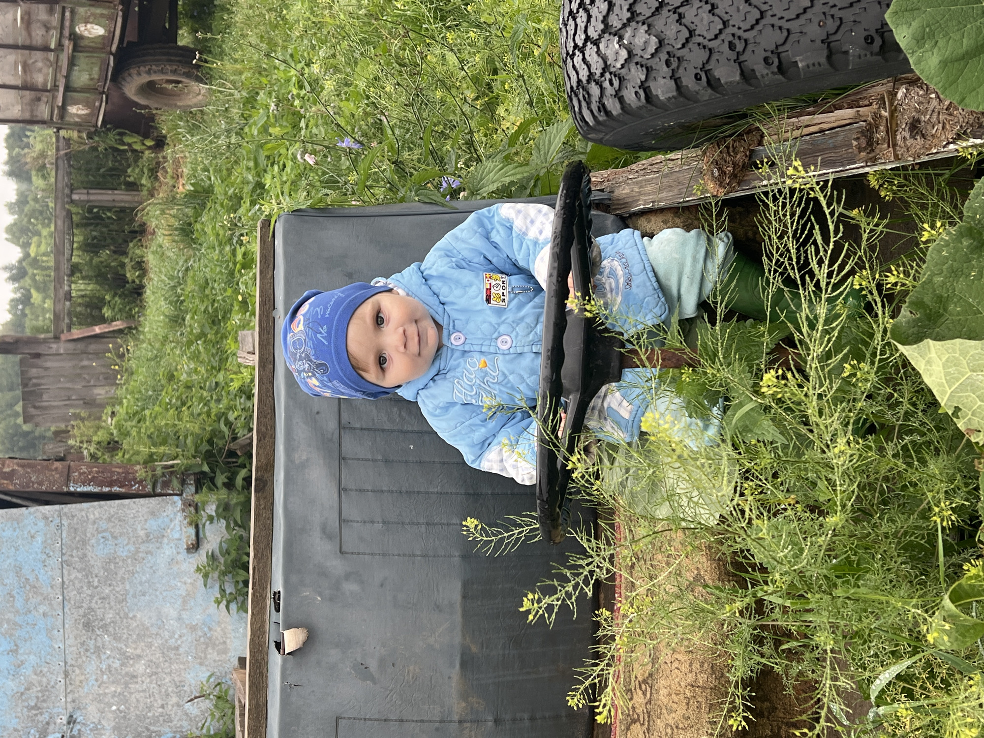 A young child, approximately one year old, sits in the driver's seat of what appears to be a small, old farm vehicle. The child wears a blue quilted jacket and a matching blue beanie with a cartoon print. They are seated on a dark gray, padded surface, with a black steering wheel in front of them. The surrounding environment is overgrown with green vegetation, including small yellow wildflowers. In the background, a rustic wooden fence and a part of a green trailer can be seen. The scene suggests a rural setting, possibly in Panteleevka, Russia, given the presence of agricultural equipment and the natural landscape. The overall impression is of a candid, everyday moment in a village.