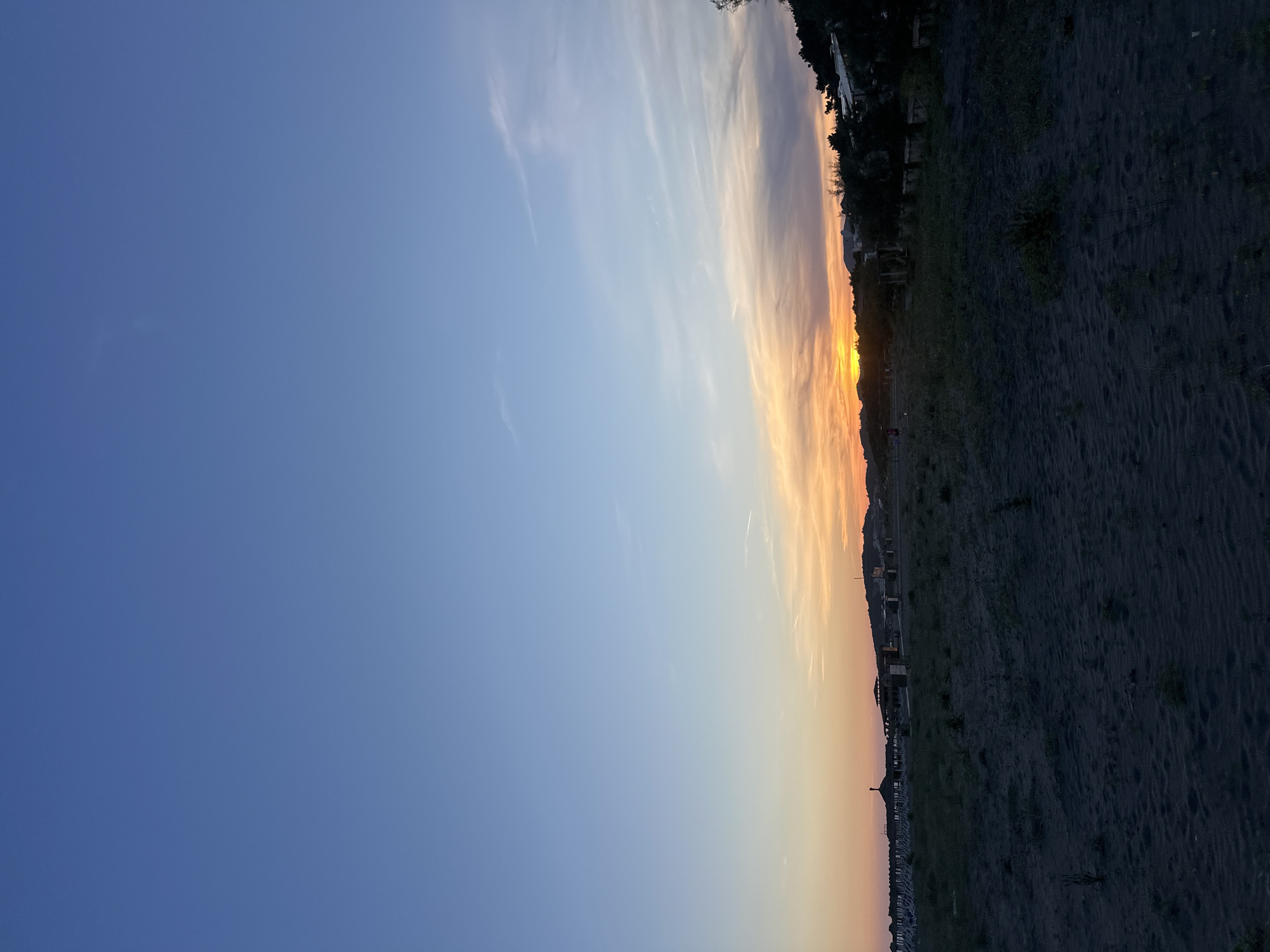 The image shows a sunset view in Donji Štoj, Montenegro. The sky transitions from a deep blue on the left to a golden orange near the horizon. Wisps of clouds are visible. The ground is a dark color. Along the horizon, there are structures resembling houses or buildings. The scene appears calm and serene, capturing the natural beauty of the coastal setting.