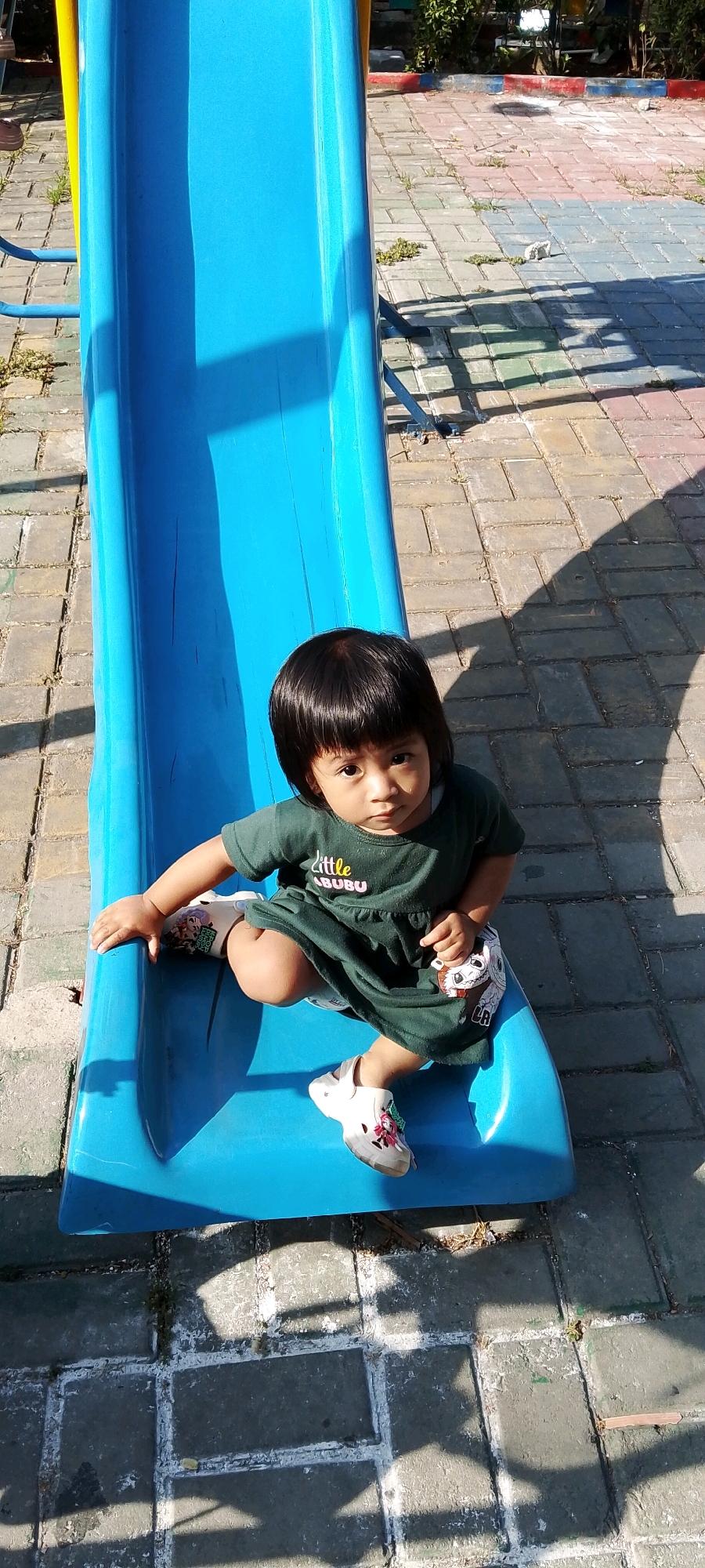 A young child sits on a blue plastic slide at a playground in Satriamekar, Indonesia. The child, who appears to be of East Asian descent, has dark hair and is wearing a dark green dress with a pink "Little BUBU" graphic on the front. The child's feet are clad in white sandals with cartoon designs. The slide is positioned on a paved area made of interlocking bricks in varying shades of gray, pink, and blue. Sunlight casts strong shadows across the playground, indicating a sunny day. The background shows foliage and parts of other playground equipment. The overall scene depicts a child enjoying a moment at an outdoor play area.
