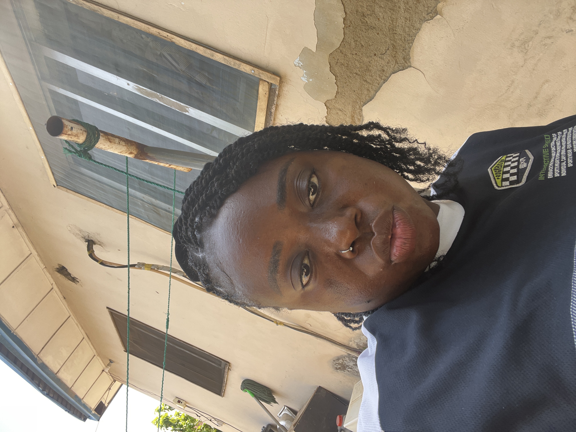 In Galadimawa Village, Nigeria, a close-up, selfie-style photograph captures a Black woman posing outdoors. She is positioned in the foreground, with a weathered, beige building serving as the backdrop. The building features a window with a wooden frame and mesh screening, and a rusty metal pipe secured with green rope. To the left of the frame, a mop with a green head leans against the wall, near a container and a red-capped bottle. The woman has dark, braided hair styled into a bun with loose braids framing her face. She is wearing a dark, long-sleeved shirt with white and black accents on the collar and a patch on the left side of her chest. She has a nose ring and her lips are slightly parted. The image is taken during daylight, indicated by the strong shadows and bright lighting.