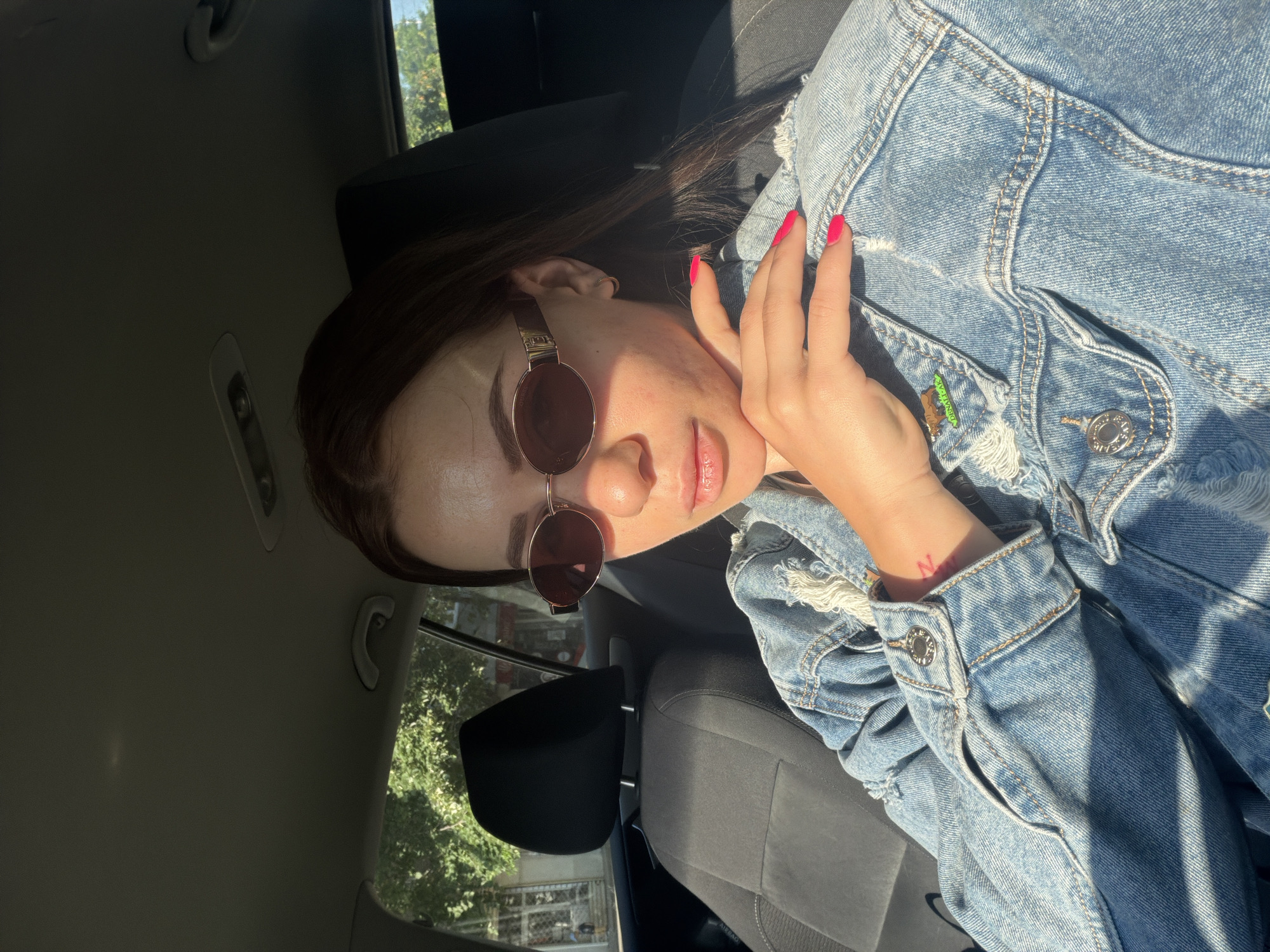 A woman with dark hair and sunglasses sits in a car. She is wearing a denim jacket with ripped details and red nail polish. Her hand is near her face. A partial view of the car's interior is visible, including the ceiling, seats, and door. Through the car window, trees and a building can be seen. However, the image does not provide enough information to determine the exact location in Plovdiv, Bulgaria.