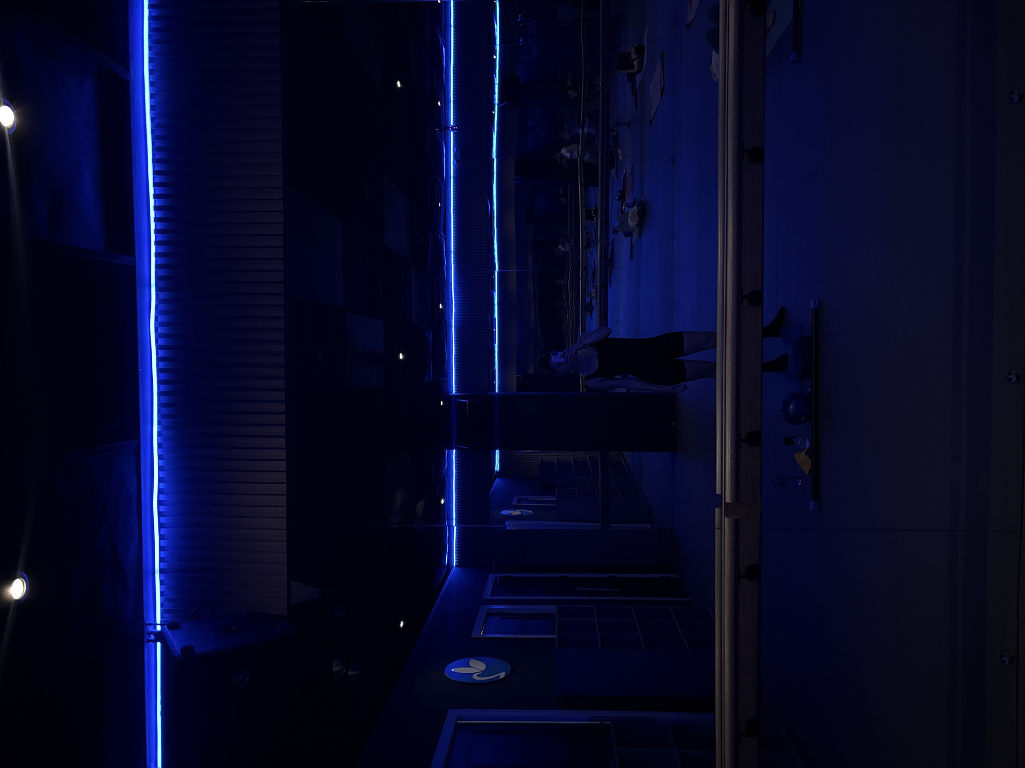 A yoga studio in Mexico City is captured in a photograph. The studio has dark blue walls and is illuminated by two vertical LED light strips, one white and one blue. The image shows a mirrored wall reflecting the yoga practitioners, mats, and general contents of the studio. People are visible in the mirror's reflection, lying on yoga mats, and appear to be engaged in a yoga class. A small circular logo with a stylized design of a leaf or petals is visible on one of the studio's lockers.