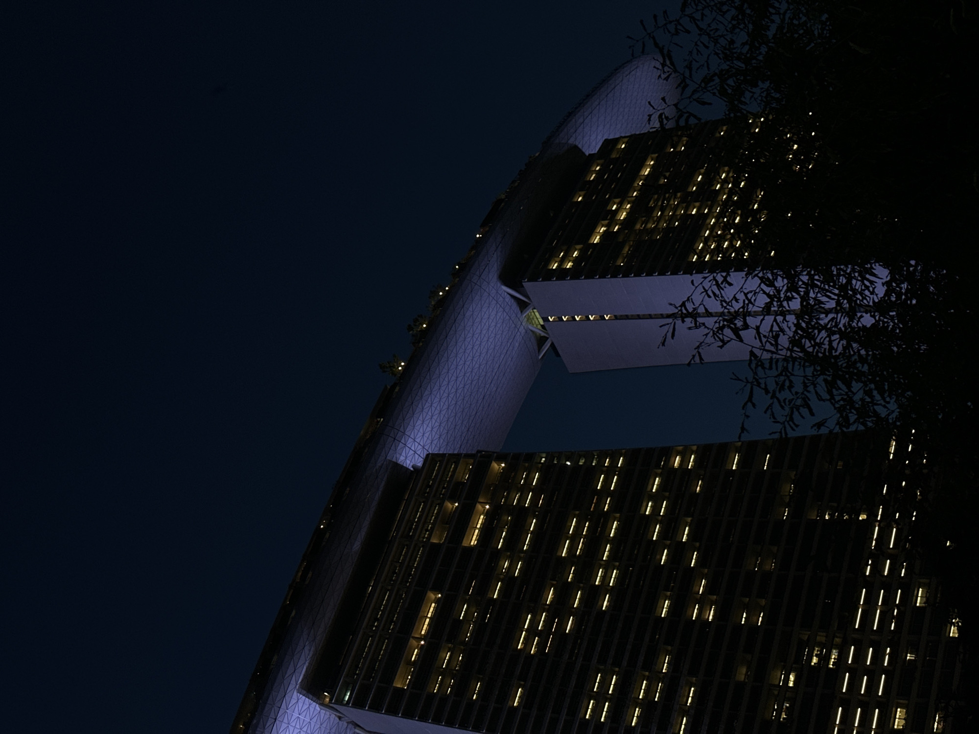 This is a low-angle, nighttime photograph of the Marina Bay Sands hotel in Singapore. The iconic building, known for its three towers topped by a Skypark, is captured against a dark blue sky. The windows of the hotel towers are illuminated with warm yellow light, creating a grid pattern that stands out against the dark facade. The curved, illuminated Skypark stretches across the tops of the towers, glowing with a soft purple hue. Silhouetted trees in the foreground partially obscure the view of the lower portions of the building, adding depth to the scene. The overall impression is one of architectural grandeur and nighttime illumination.