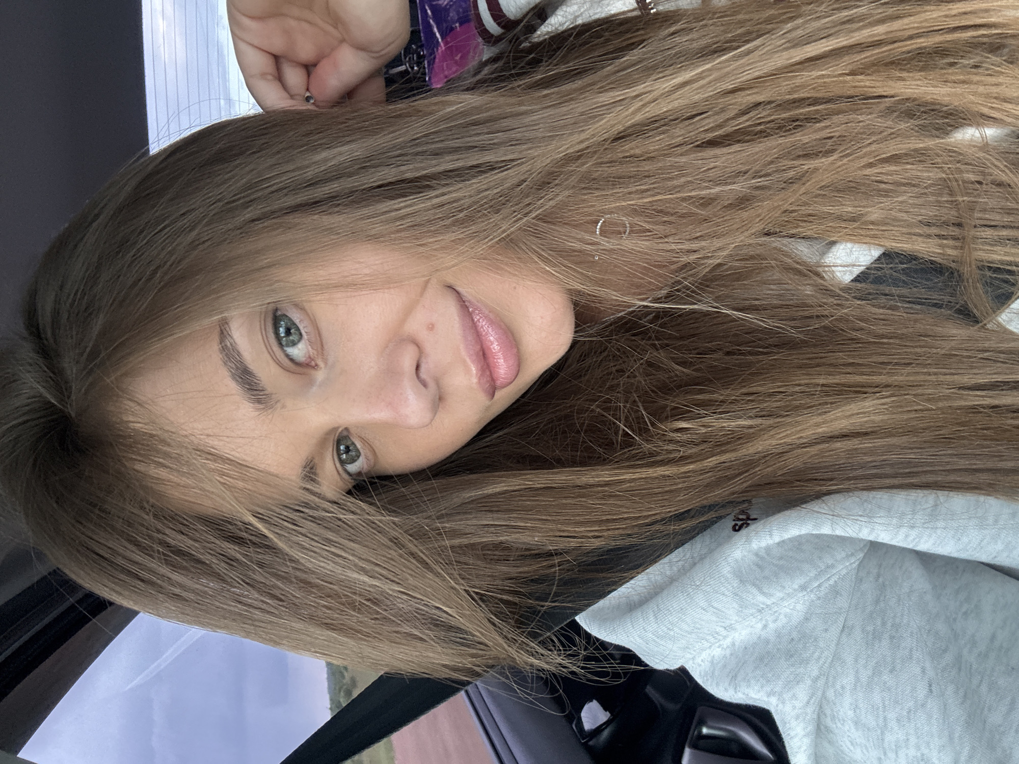 The image is a close-up, selfie-style photograph of a young woman with long, light brown hair, taken from the chest up. She is seated in what appears to be a vehicle, with the car's interior visible on the left side of the frame, including a seatbelt buckle and part of the door. The woman is looking towards the camera with a slight smile. Her makeup is subtle, with emphasis on her lips and eyes. She is wearing a light-colored hooded sweatshirt. The background outside the car window suggests an outdoor, possibly rural or semi-rural, setting with a cloudy sky. The image does not contain any specific landmarks or indicators to pinpoint a location within Kuyeda, Russia.