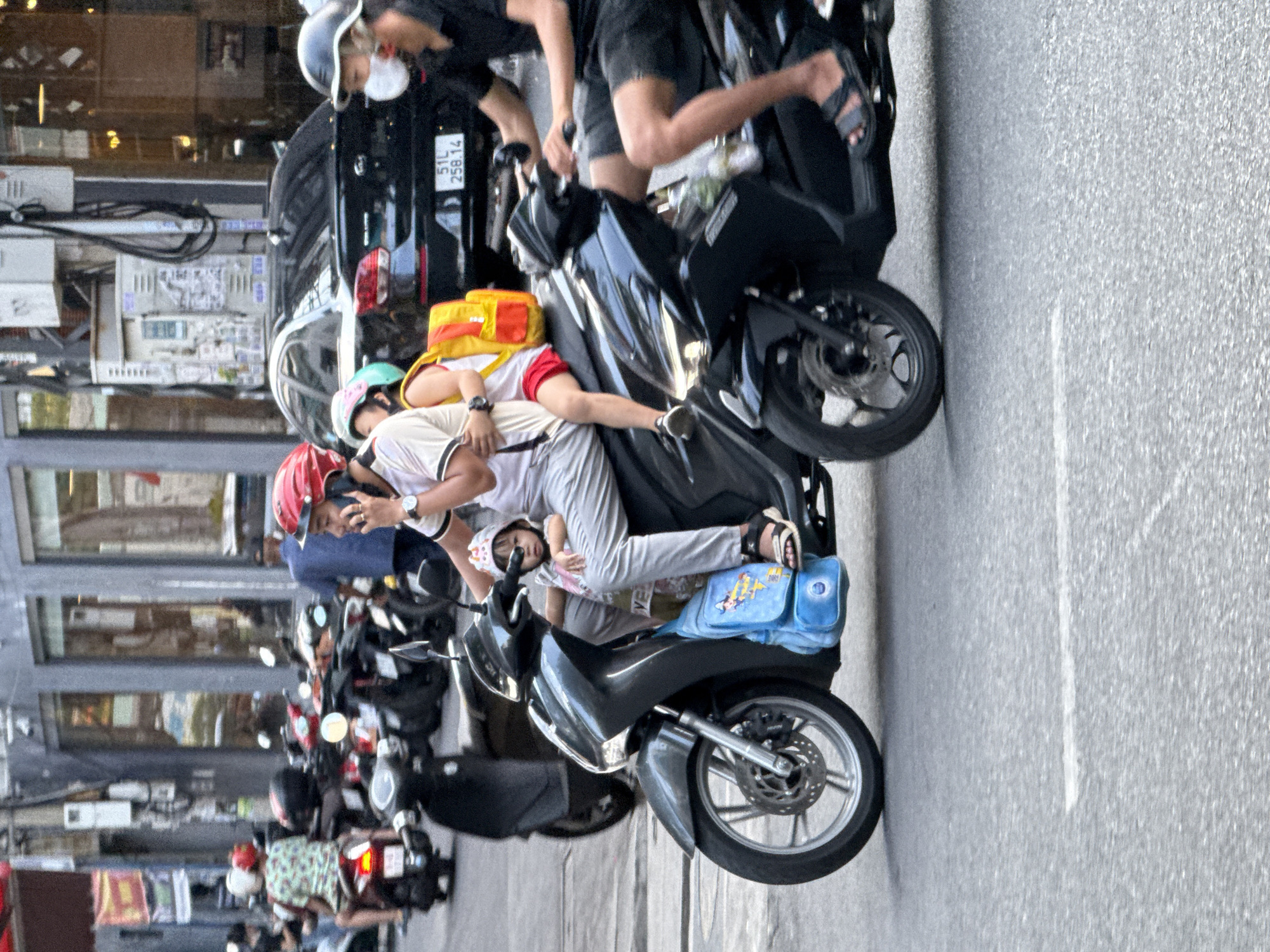 The image depicts a street scene in Nha Trang Ward, Vietnam, featuring a family riding on a motorcycle. A man, wearing a red helmet and a light-colored shirt, is driving the motorcycle while speaking on his phone. He has a watch on his left wrist. Next to him, a woman is seated, wearing a pink top and red shorts, with a yellow backpack strapped to her back. A young child, wearing a white and pink outfit and a light-colored helmet, is seated between the two adults. In the foreground, a blue backpack with a cartoon character is visible. Another motorcycle, ridden by a person wearing a black shirt and a white face mask, is to the right. The background shows buildings, parked motorcycles, and a black car with a license plate displaying "51L 258.14". The scene appears to be on a paved street, suggesting a public urban environment.