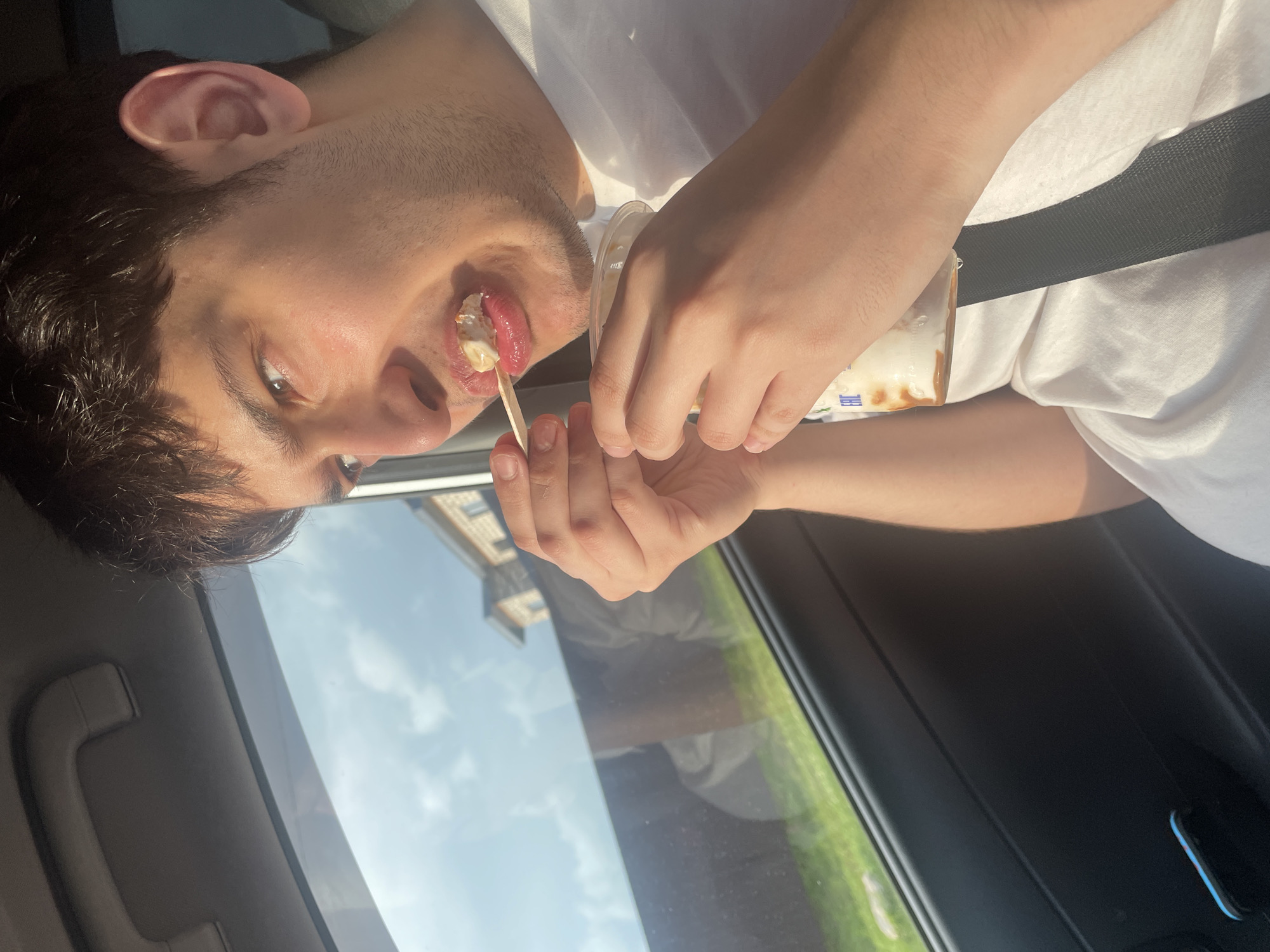 A young man is shown from the chest up, inside a vehicle, looking out the passenger-side window. He is holding a small wooden spoon with food on it and bringing it to his mouth. He is wearing a white t-shirt. Outside the window, green grass and a building are visible, suggesting a suburban or rural setting, potentially in Gavrilovka, Russia. The interior of the car is dark, with the window reflecting the sky.
