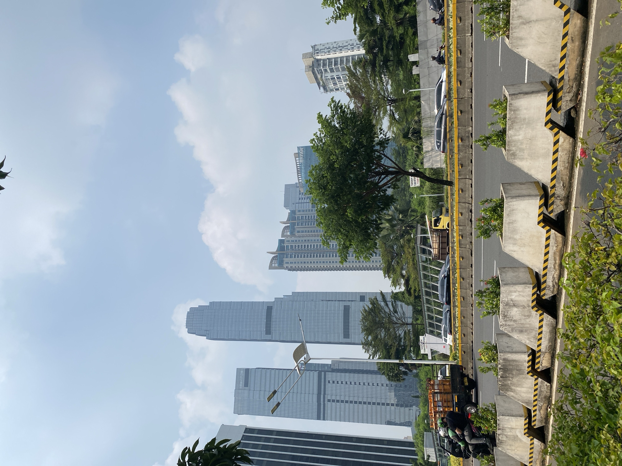 The image depicts a street view in Central Jakarta, Indonesia, on a bright, clear day with scattered clouds. The scene features a multi-lane road with traffic, including motorcycles, cars, and a truck. In the foreground, there are concrete barriers with yellow and black hazard stripes, planted with small green bushes. A motorcycle carrying two people is moving on the road, with the passenger holding onto a load of what appears to be orange crates.
The midground reveals a row of trees and greenery separating the road from an elevated walkway or bridge. Behind the trees, several modern high-rise buildings dominate the skyline. Two prominent, sleek skyscrapers with glass facades are positioned centrally, with other office or residential buildings of varying heights and architectural styles surrounding them. A street lamp with multiple lights stands tall near the center of the image.
The background is dominated by the clear blue sky with white clouds. The overall impression is of a bustling urban environment in a major city.