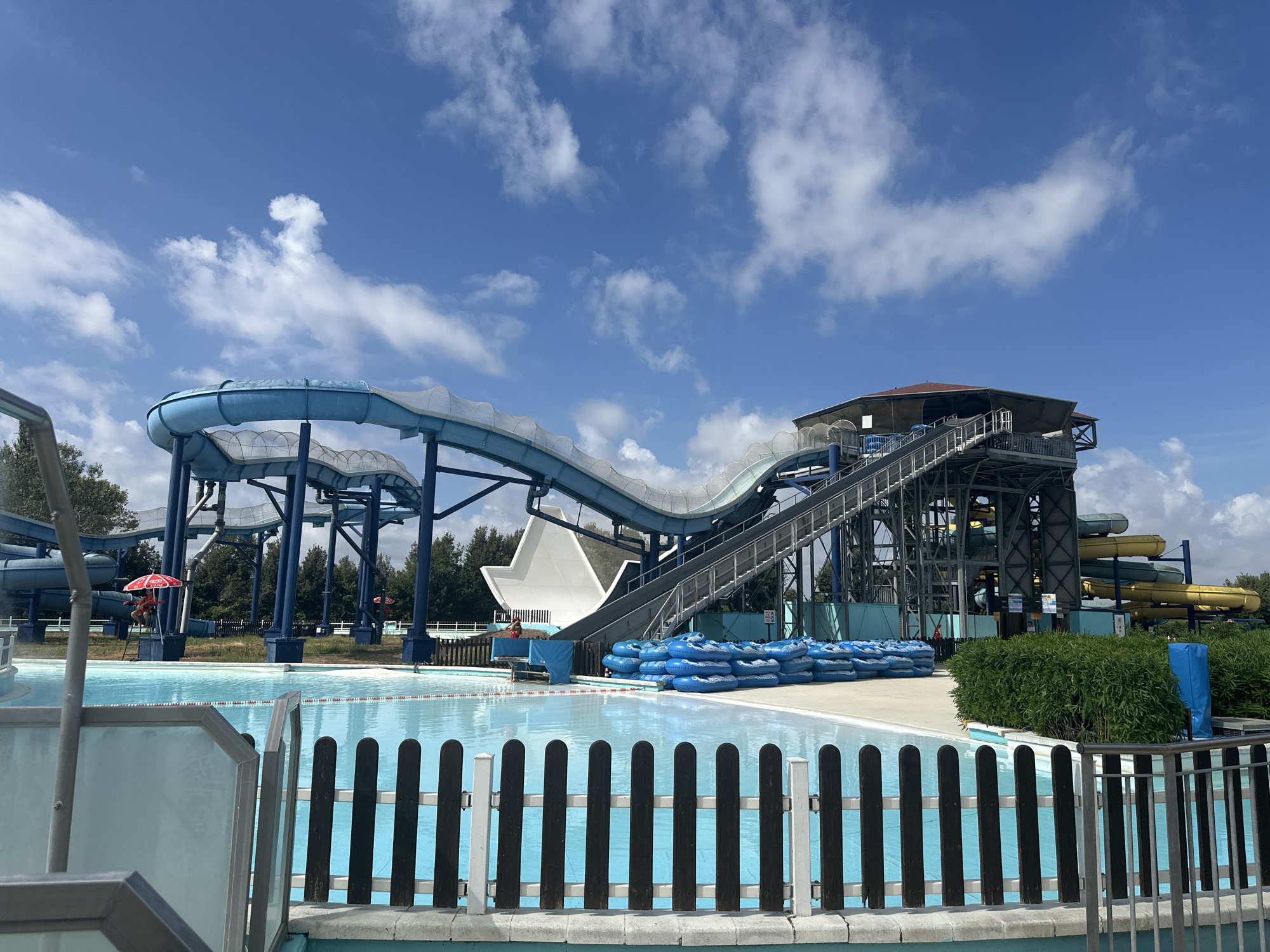 The image captures a section of a water park under a bright blue sky dotted with white clouds. In the foreground, a shallow, turquoise swimming pool is partially visible, bordered by a black and white picket fence. Beyond the pool, a large blue water slide structure dominates the scene, with its winding tubes supported by a dark metal framework. At the base of the slide, a stack of blue inflatable rings is neatly arranged. To the right, a dense green hedge runs along the edge of the park. The background reveals more water slides in yellow and green, and beyond that, a line of trees. A lifeguard wearing a red uniform and holding a red umbrella stands near the pool. The overall impression is that of a sunny day at a recreational facility. The city of Ravenna, Italy, is known for its rich history and cultural heritage, but this image depicts a modern water park which is unlikely to be located within the historic city center.