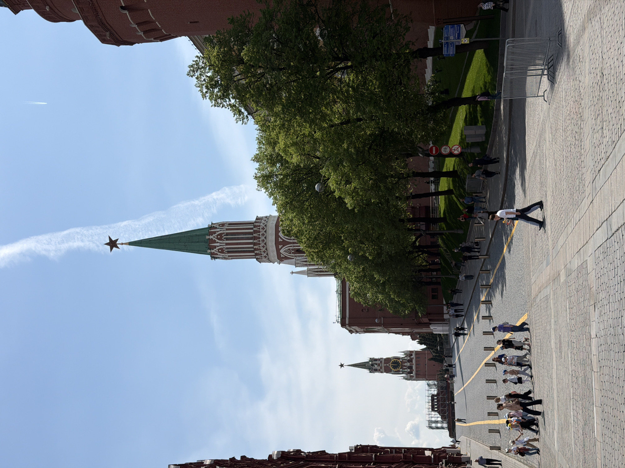 The image captures a view of Red Square in Moscow, Russia. Several of the Kremlin's towers are visible, including one topped with a star and another with a clock face. Trees line the square, and people are walking along a path marked by yellow lines on the cobblestone pavement. A contrail is visible in the sky. Part of the red brick wall of the Kremlin is present at the top and bottom of the image.