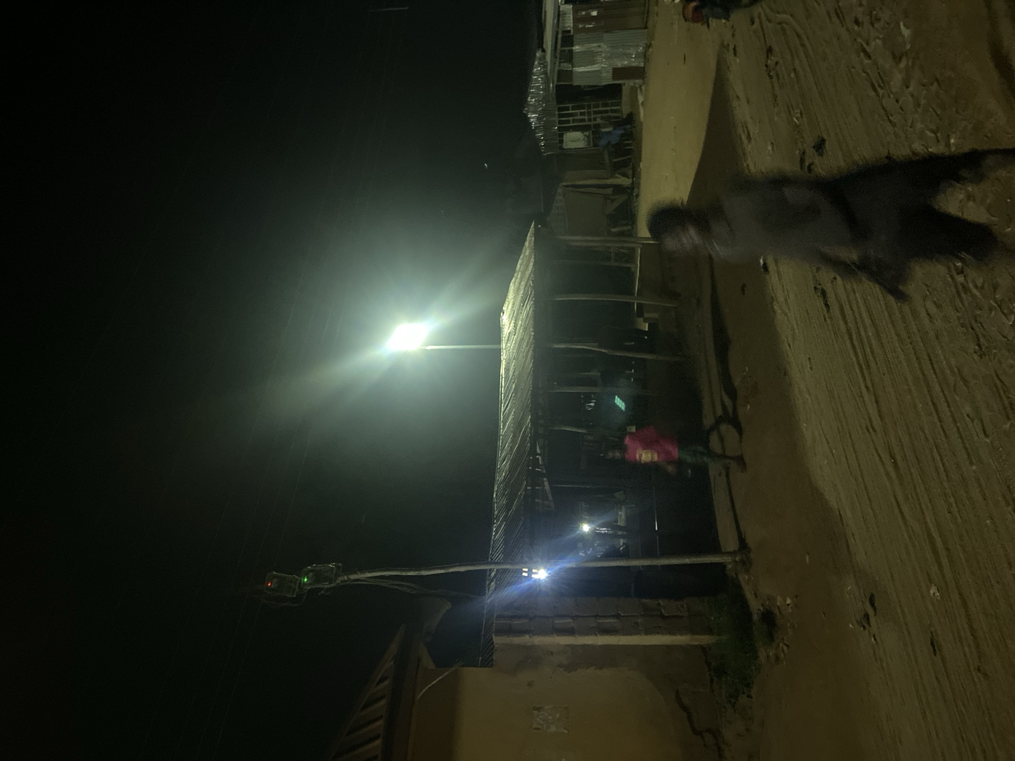 The image depicts a dimly lit nighttime scene in Kuson, Nigeria. A street is illuminated by a bright overhead lamp, casting a strong glow and creating lens flare. To the left, a traffic signal is visible with a green light illuminated. Adjacent to the road are structures that appear to be makeshift buildings with corrugated metal roofing. A person, partially obscured and blurred, is walking on the sandy ground on the right side of the image. Another person, wearing a pink garment, is visible near the buildings. The ground appears to be dirt or sand, suggesting an unpaved road. The overall impression is of a simple, possibly rural or semi-urban, street at night, with limited artificial lighting. There is no visible text in the image.