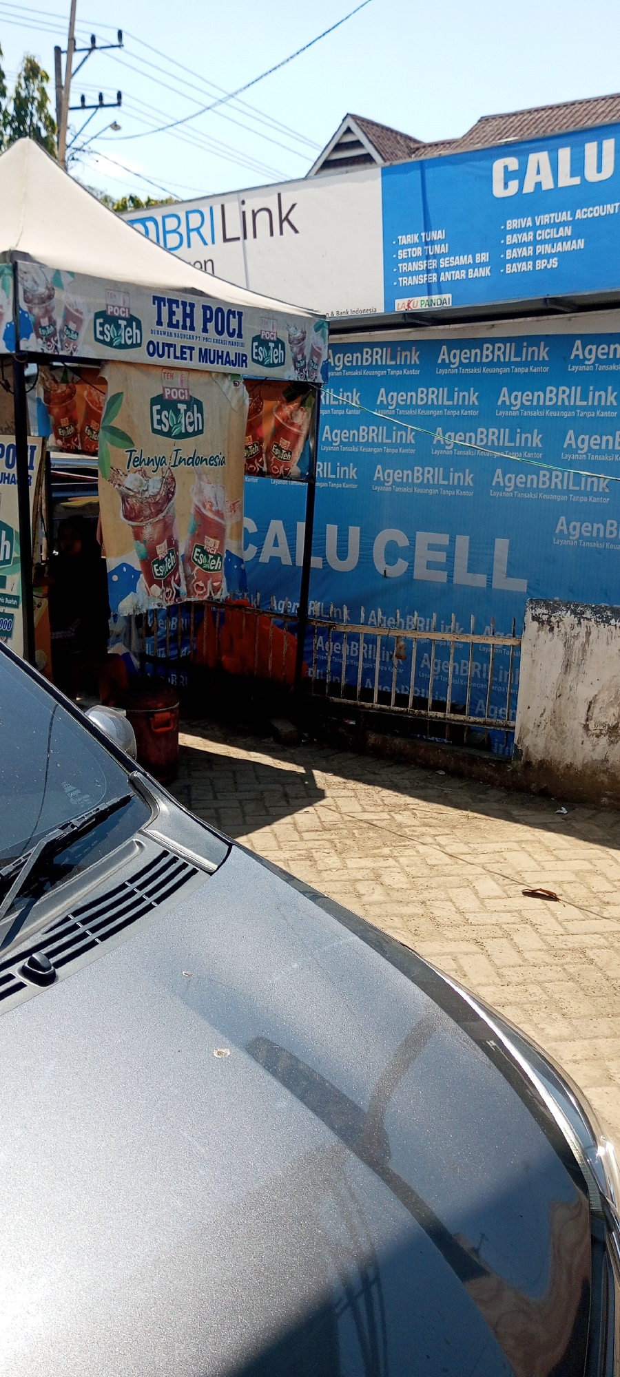 In Lempa, Indonesia, a street-side stall operates under the bright sun. A white tent covers the stall, displaying advertisements for "TEH POCI Eso Teh" and "BRI Link". Behind the stall, a blue banner promotes "CALU CELL" and lists financial services such as cash withdrawal, bill payments, and bank transfers. The stall appears to be an agent for BRI Link, a financial service from Bank Rakyat Indonesia. A gray car is parked in the foreground, its hood reflecting the sunlight and the surrounding environment. The ground is paved with bricks, and a few fallen leaves are visible. The overall scene suggests a typical day of commerce in a local Indonesian town, with a blend of food and financial services offered to passersby.