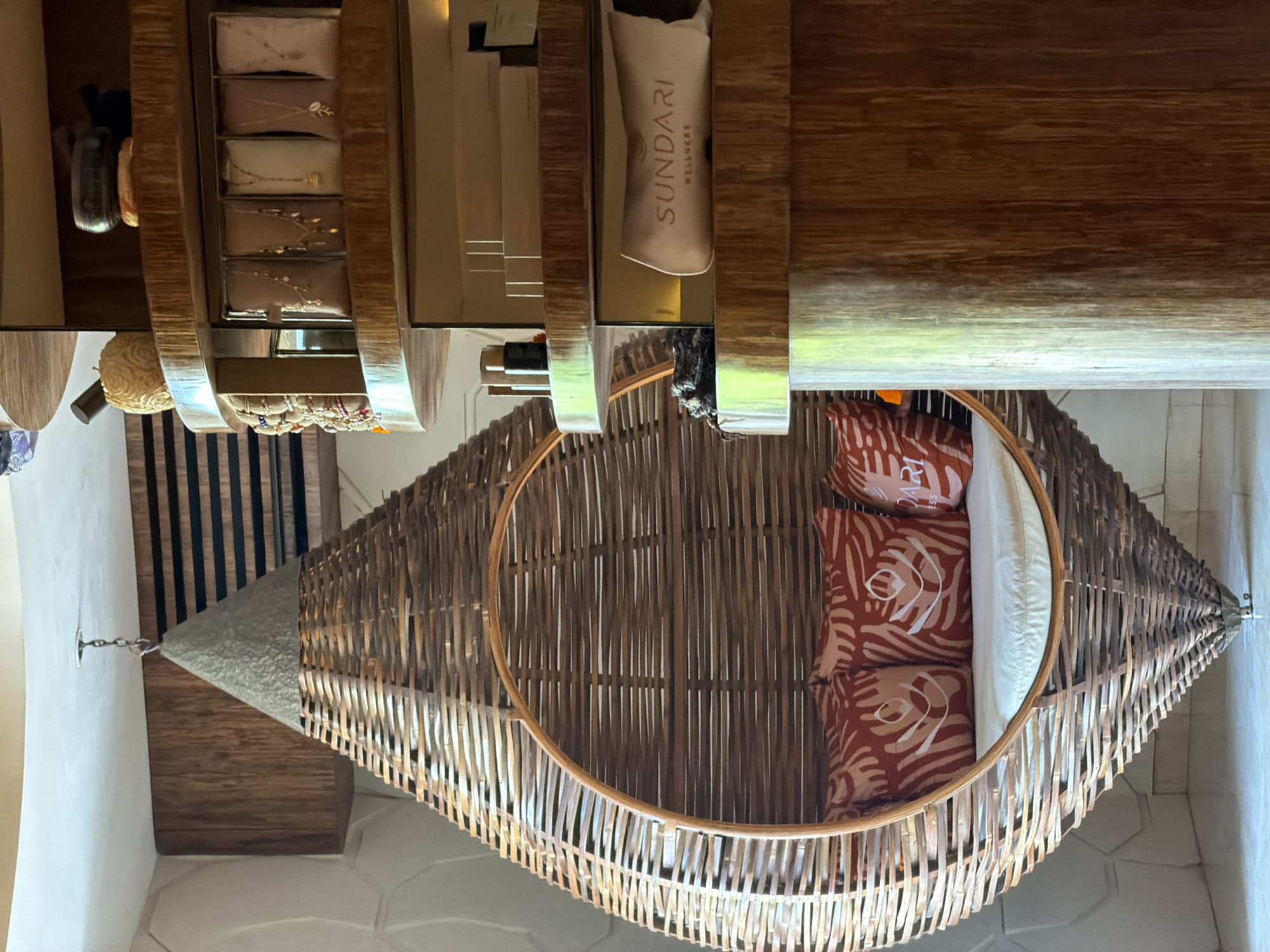 The image captures an elegantly designed indoor space, likely a wellness center, spa, or resort lounge, located in North Kuta, Indonesia. The dominant feature is a large, suspended oval-shaped chair or daybed, meticulously crafted from interwoven natural materials like bamboo or wicker, giving it a rustic yet sophisticated appearance. Inside the hanging chair, several plush pillows are arranged for comfort: three reddish-orange pillows with a subtle, light-colored organic pattern, and one plain white cushion.
In the background, above the chair, a display unit with wooden and metallic accents showcases various items. On an upper shelf, several rectangular, light-toned packages, possibly soaps or small gifts, are neatly arranged, some featuring delicate designs like leaves or branches. Below these, a smaller stand holds multiple beaded bracelets, suggesting a retail or gifting area. Further to the right on a shelf, a cream-colored pouch is visible, bearing the text "SUNDARI WELLNESS" in a refined font, identifying a brand likely associated with spa or health products.
The interior design emphasizes natural textures and a warm color palette, with visible wooden elements and light-colored walls, contributing to a serene and inviting ambiance. The scene is devoid of people, allowing the focus to remain on the luxurious and comfortable furnishings, hinting at a space dedicated to relaxation and well-being. The lighting appears to be ambient indoor illumination.