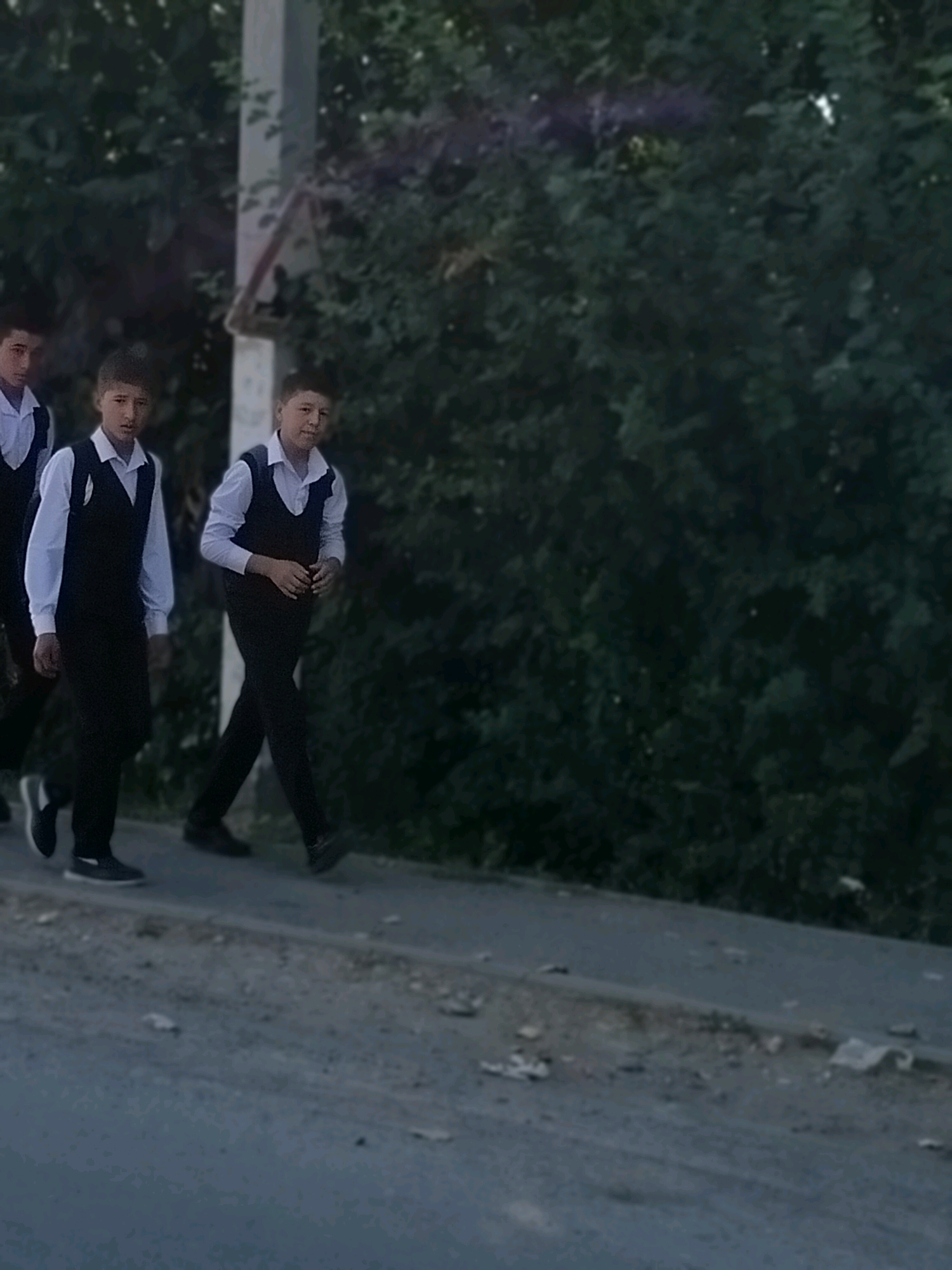The image shows three young boys, appearing to be students, walking on a paved path next to a road. They are all wearing similar school uniforms consisting of a white shirt and a dark vest. The boy in the center is looking directly at the camera, while the other two are looking forward. The background is dominated by dense green foliage, likely trees or bushes, with a utility pole visible to the left of the boys. The ground in front of them is a mixture of asphalt and dirt, with some scattered debris. The lighting suggests it is either early morning or late afternoon. The location is identified as Көлкент, Kazakhstan.