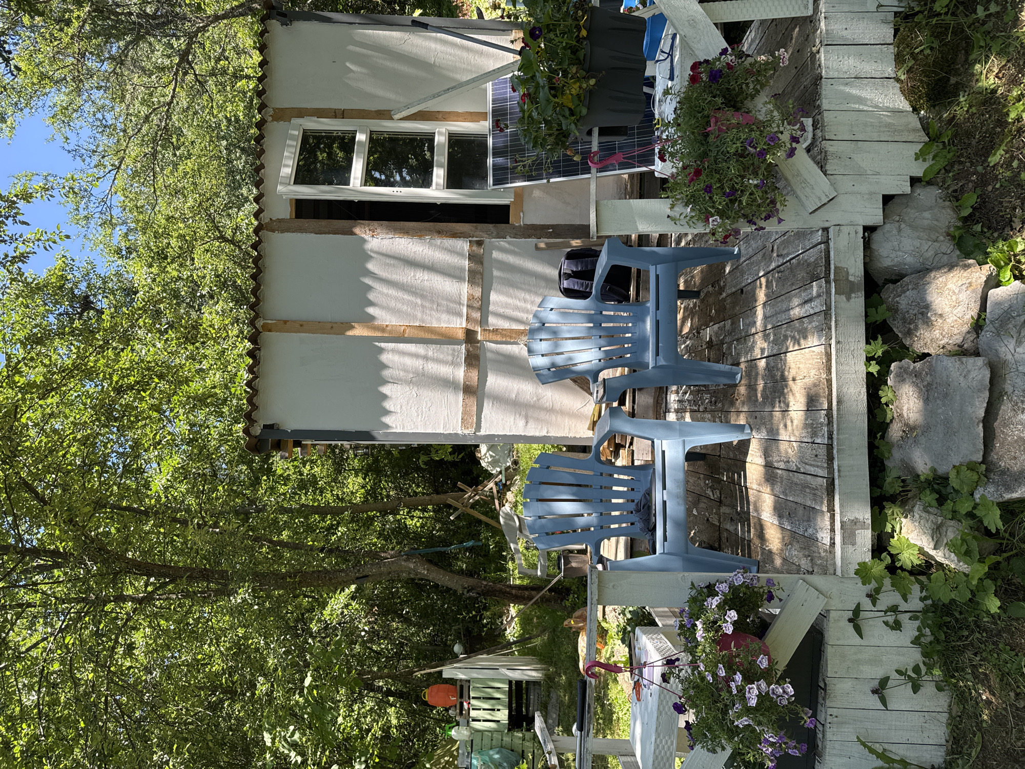 The image depicts an outdoor scene in Dramsha, Bulgaria, featuring a small, unfinished structure with a corrugated roof and white walls. Two blue plastic chairs are placed on a wooden deck in front of the structure. Hanging baskets filled with flowering plants are attached to the railing of the deck. To the right of the structure, a solar panel and more potted plants are visible. In the background, lush green trees and foliage create a natural setting. There are no people present in the image.