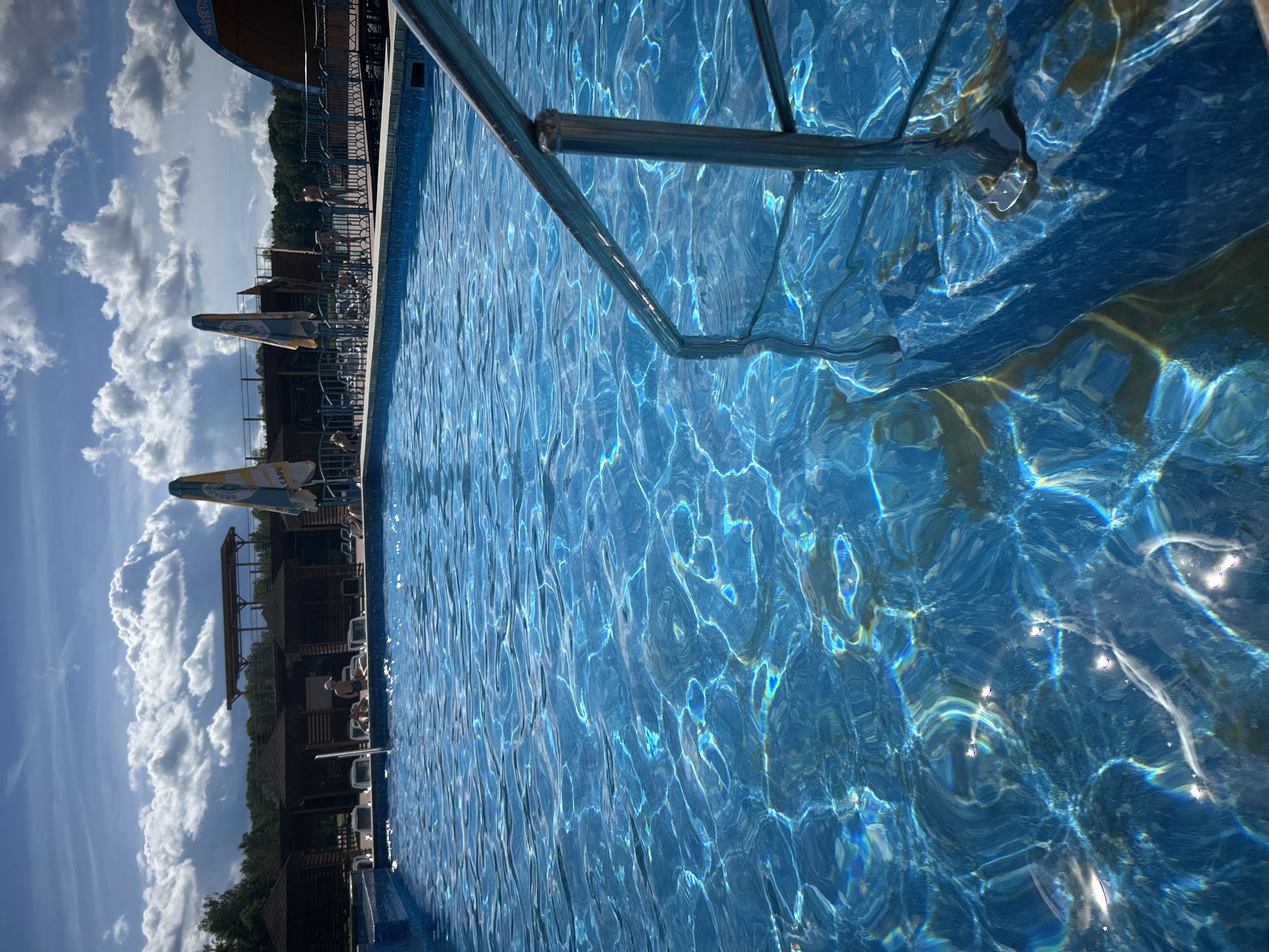 The image shows a swimming pool in the city of Yubileynyy, Russia. The pool is filled with clear blue water, which is reflecting the sunlight and creating shimmering patterns on the surface. A metal handrail extends into the pool from the right side of the frame. In the background, there are several wooden gazebos with thatched roofs, lounge chairs, and umbrellas. A few people can be seen relaxing by the pool. The sky is cloudy with patches of blue sky visible. The overall impression is one of a relaxing summer day at a resort.