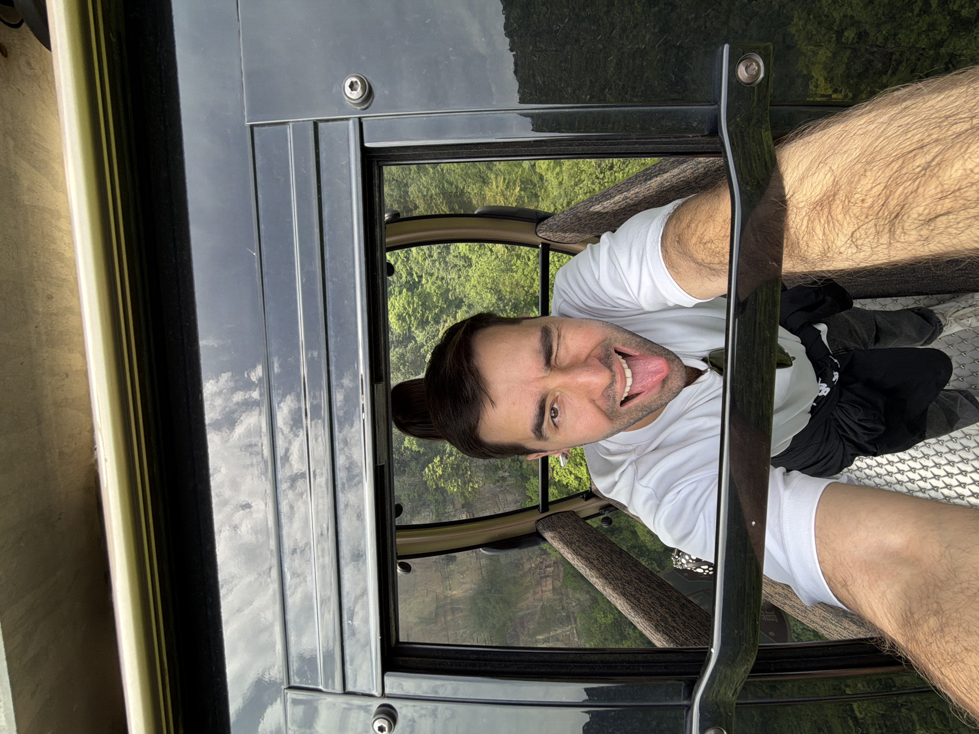 A man is inside a gondola with a view of the forested landscape of Wulingyuan, China, sticking his tongue out and winking. The man is wearing a white T-shirt. His legs are visible. A pair of black pants lie on the gondola floor. The view through the window shows trees and rock formations. The gondola car is black.