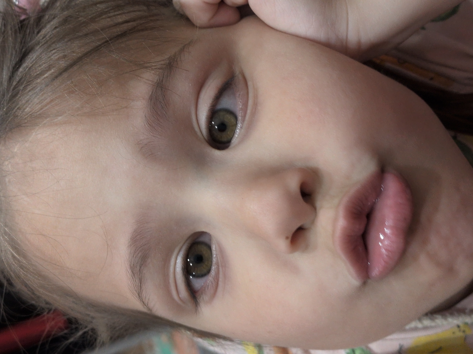 This close-up portrait captures the face of a young child, likely a girl, tilted diagonally within the frame. The child has striking green-brown eyes that gaze directly at the viewer. Her lips are playfully pursed in a "fish face" or pout. Light brown hair frames her face, appearing soft and fine. Her skin is fair and smooth. A small hand, possibly her own, is partially visible at the top of the frame, resting gently near her forehead.
The background is soft and out of focus, showing indistinct patterns and colors of light-colored fabric, suggesting an indoor setting, perhaps on a bed or sofa. The child's expression conveys a sense of innocence and playfulness. There are no specific objects other than the child herself, nor any visible text. The image does not contain any visual cues to confirm the provided location context of Sochi, Russia.