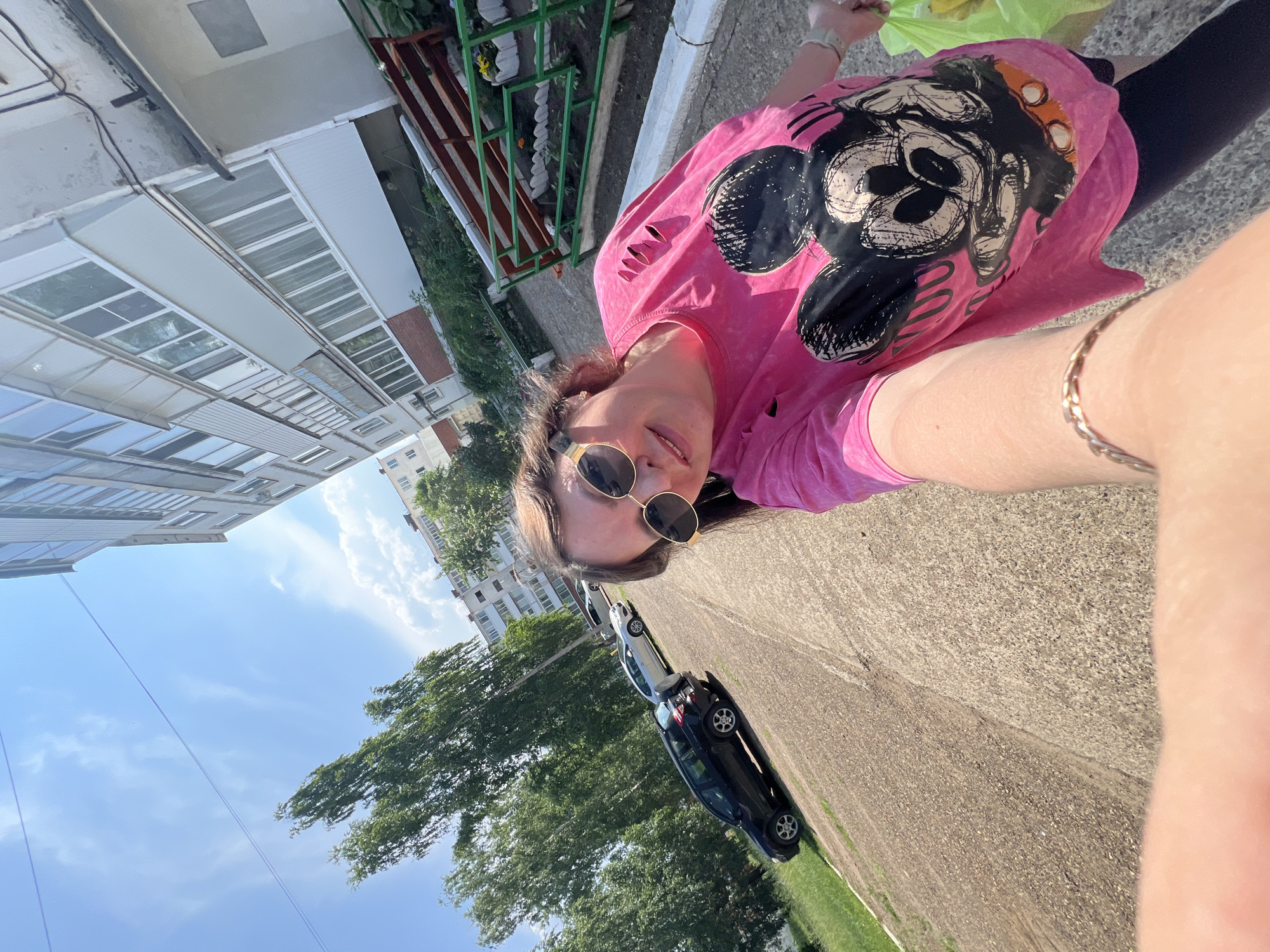 A woman takes a selfie outside on a sunny day. She is wearing a pink t-shirt with a large graphic of Mickey Mouse's face on it and torn sleeves. She has on sunglasses and a silver bracelet. In the background, there are cars parked on a gravel lot, trees, and a multi-story building. The location is Noviy, Russia.