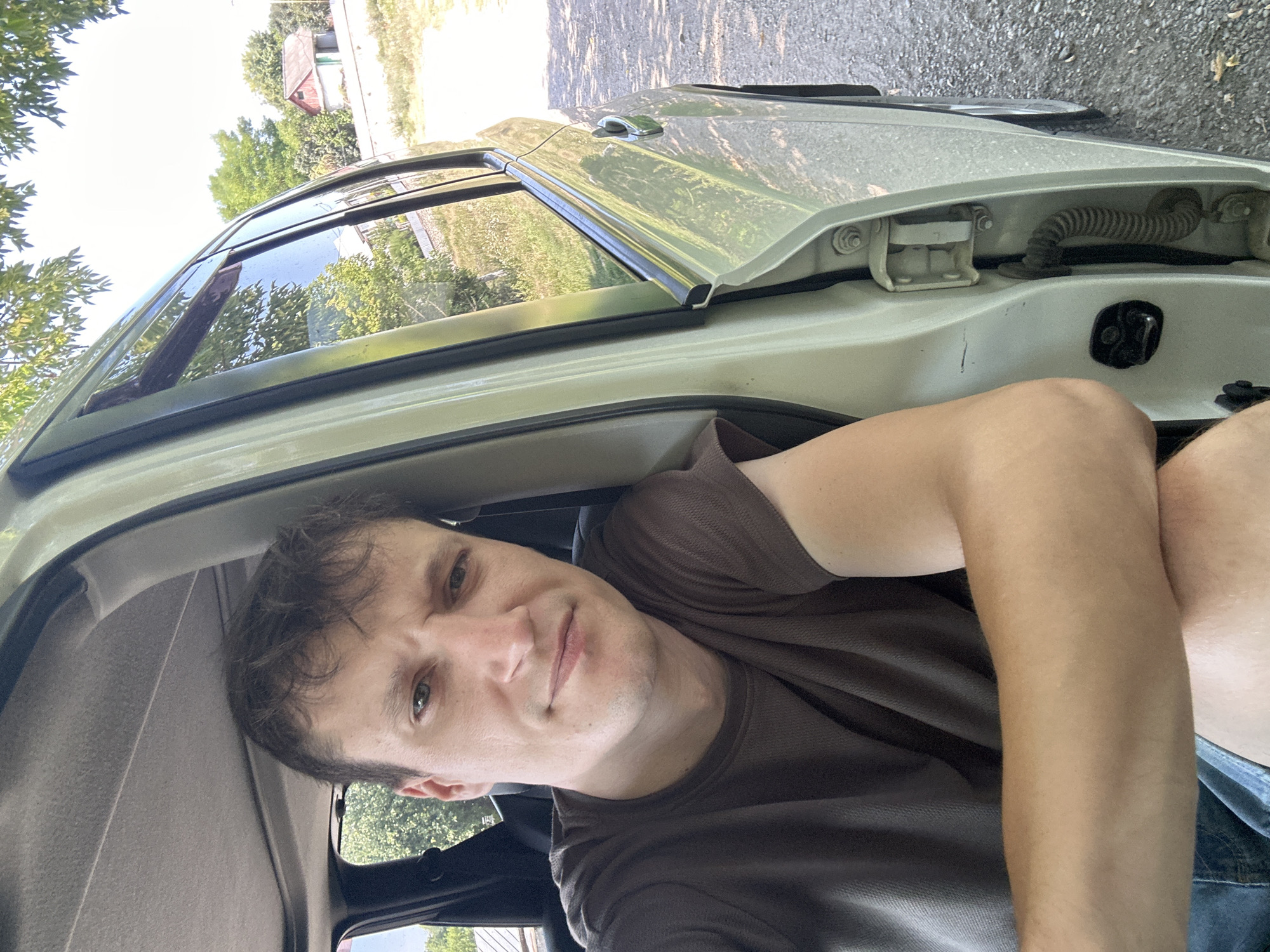 A fair-skinned male, appearing to be in his late 20s or early 30s, is taking a selfie from inside a light-colored vehicle. He is wearing a dark brown t-shirt and denim shorts. His left arm is resting on his lap, and his right arm is extended, holding the camera. He has short, dark brown hair and is looking directly at the camera with a neutral expression.
The vehicle's interior is visible, including the sunroof and part of the dashboard. The driver's side door is open, revealing the exterior environment. The background suggests a rural or semi-rural setting, with trees, grass, and a small building with a red roof visible in the distance. The ground outside the car appears to be gravel or dirt, with fallen leaves.
The location is identified as Makiv, Ukraine, based on the provided context. The lighting suggests it is daytime, likely late morning or early afternoon, with sunlight casting shadows. There are no other individuals or notable objects in the frame besides the man and his immediate surroundings.