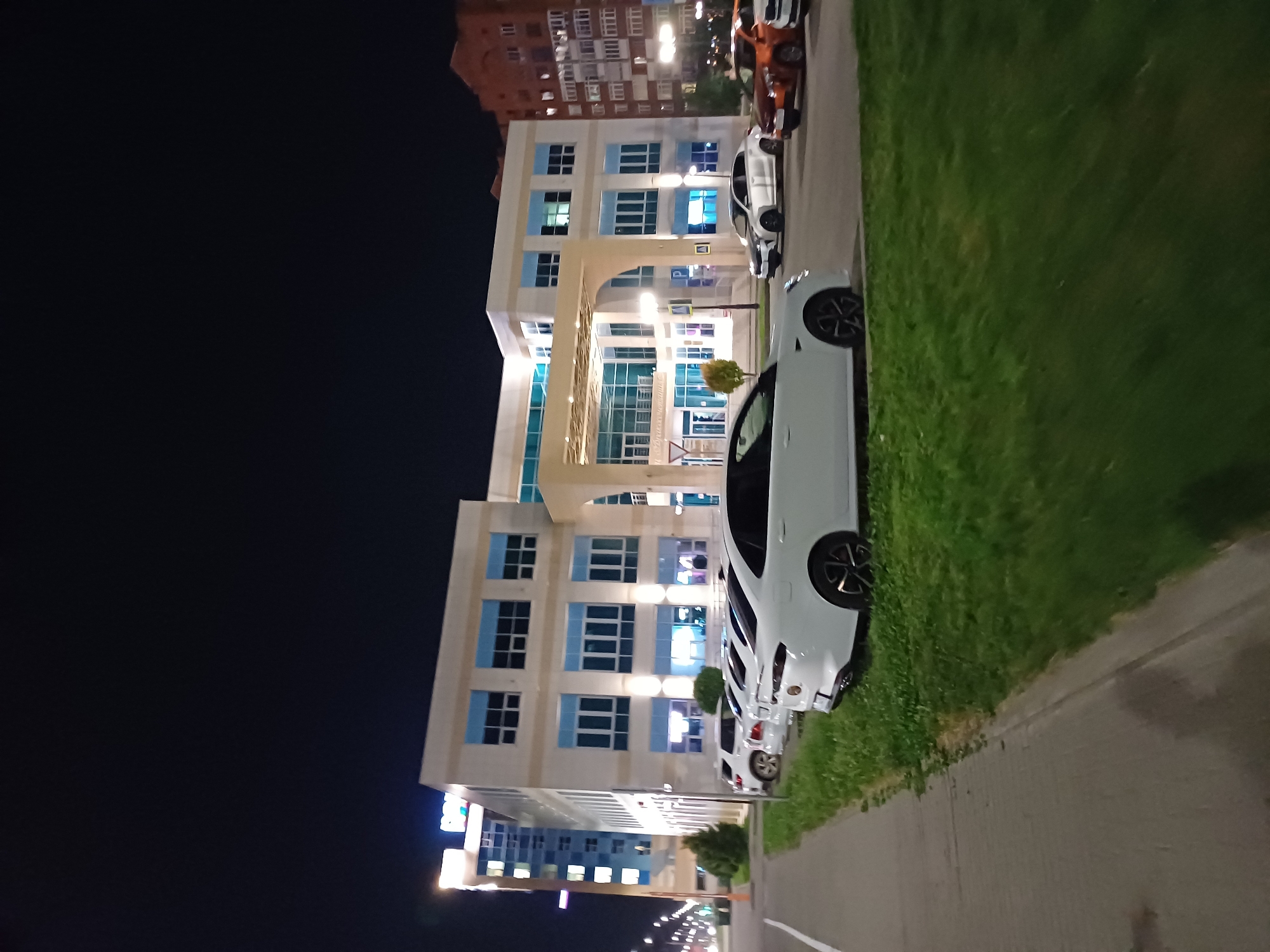 An exterior night shot of a modern office building in an urban setting. The building has a light-colored facade with large, blue-tinted windows arranged in a grid pattern. An arched entranceway is visible, leading to a covered area with patterned glass walls. Several cars are parked in front of the building, including a white sedan and an orange sedan. The area is illuminated by streetlights and lights from within the building, casting reflections on the wet pavement. A sidewalk borders a grassy area in front of the parking lot. In the background, a taller building with illuminated windows and a brick building with multiple floors are visible. The sky is dark, indicating it is nighttime.