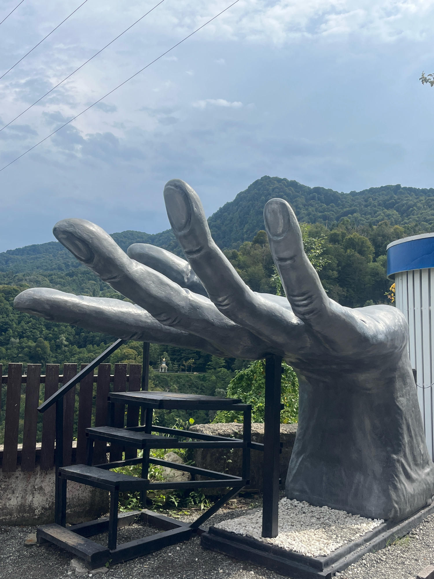 An outdoor, eye-level, medium shot shows a large, grey, open hand sculpture positioned in front of a mountainous, forested landscape. The sculpture, which appears to be made of concrete or a similar material, is the primary subject. Its fingers are extended upwards and slightly to the left, creating a sense of offering or reaching out. The surface of the sculpture has a rough texture.
To the left of the sculpture, a dark metal staircase with wooden steps is visible. It appears to be a simple, functional structure. Behind the stairs and sculpture, a wooden fence runs horizontally. Beyond the fence, a steep, heavily forested slope descends, with a small, light-colored building featuring a golden dome visible in the distance, nestled among the trees. The background is dominated by rolling hills covered in dense green trees, with a cloudy, grey sky overhead. In the upper left corner, thin, dark power lines stretch across the sky. To the right of the sculpture, a portion of a cylindrical white structure with a blue band is visible. The ground in the foreground is covered with gravel and small white stones around the base of the sculpture. There are no people visible in the image. The overall impression is a blend of natural scenery and man-made art installation. The specific location within the city of Khartsyz Pervyi, Russia, is not ascertainable from the visual information alone.