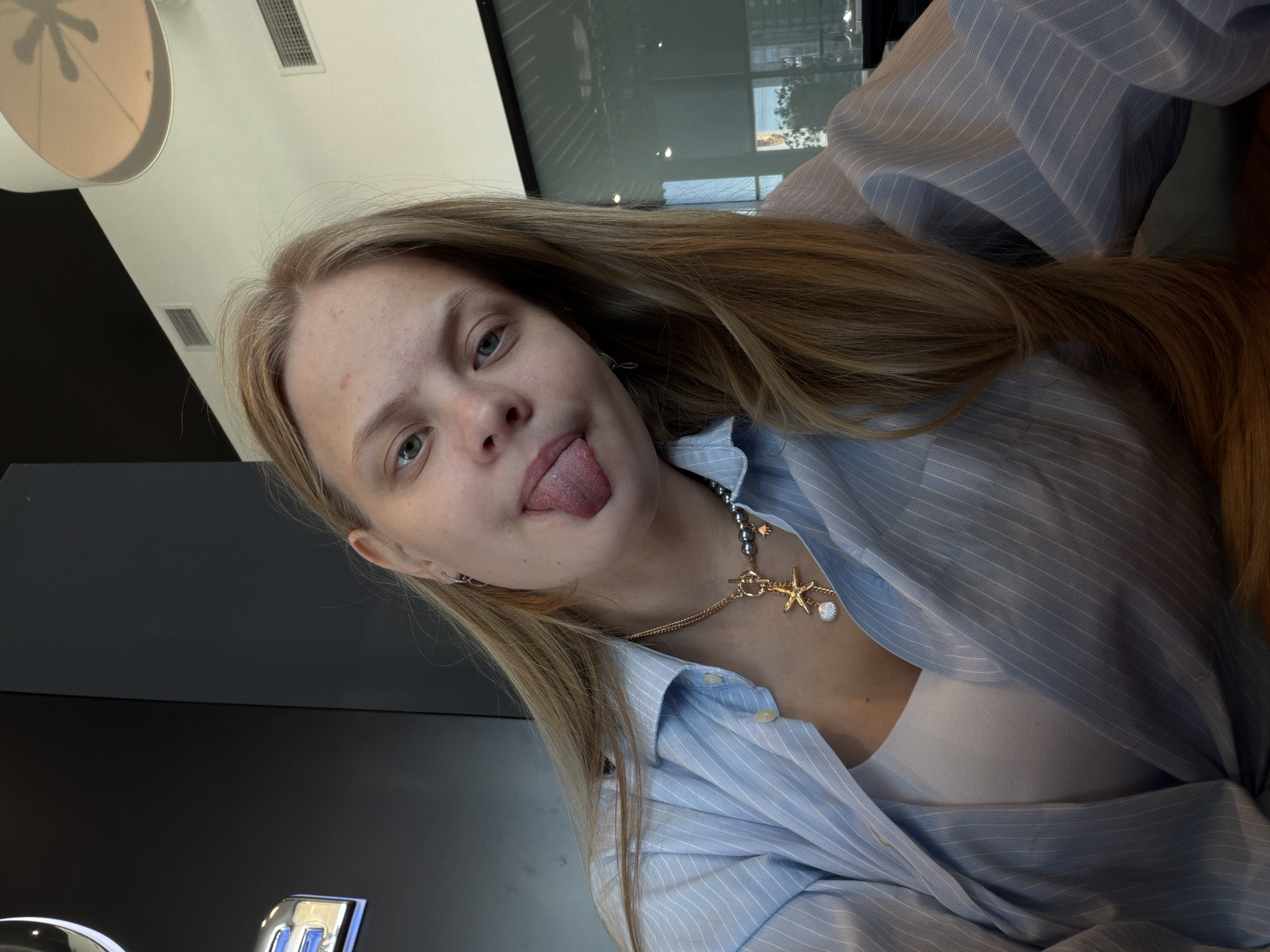 A young woman with light blonde hair is photographed indoors, sticking her tongue out towards the camera. She has light skin and noticeable blemishes on her face. She is wearing a light blue striped button-down shirt, which is open enough to reveal a white top underneath. A necklace with a starfish and seashell charm is visible around her neck.
The background suggests an interior setting. A partially visible lamp fixture is in the upper left corner. A window is in the upper background. A black object is located in the lower left corner of the image.
The location is not discernible as Izhevsk, Russia from the visual details in the image alone.