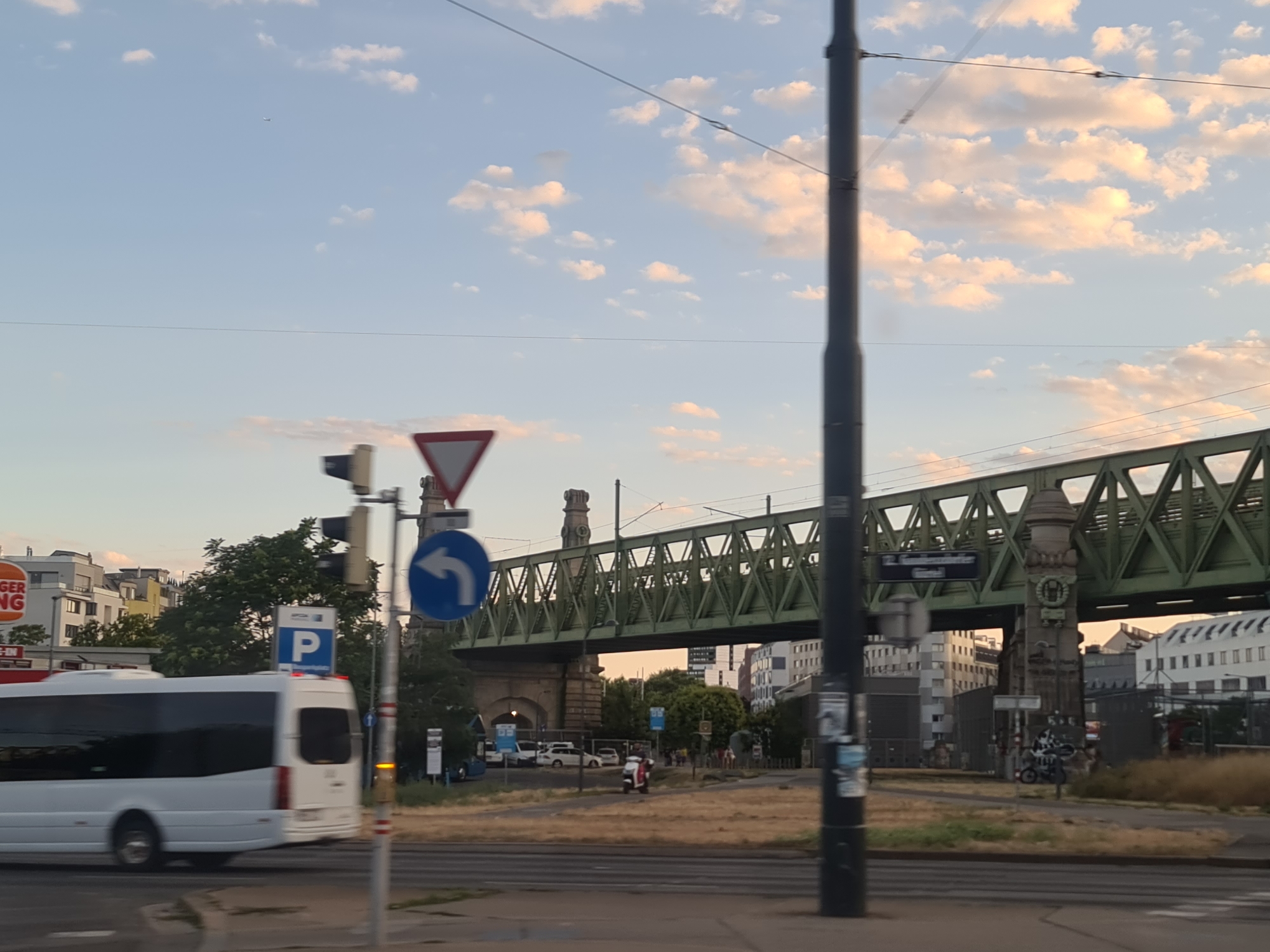 The image depicts an outdoor scene with a light blue sky filled with scattered white and pink clouds, suggesting either early morning or late afternoon. A large, green metal railway bridge dominates the middle ground, supported by stone pillars. Various buildings, including a Burger King restaurant, are visible in the background on the left. A white bus is in motion on the left side of the frame, and other vehicles, including cars and a scooter, are present on the road and in the distance. Several traffic signs are visible, including a "Give Way" sign and a blue sign with a white "P" indicating parking. A tall, dark pole, likely a lamppost or utility pole, stands prominently on the right side of the image. The overall environment appears to be urban. The scene is likely located in Vienna, Austria, based on the architectural style of the buildings and the presence of a Burger King.