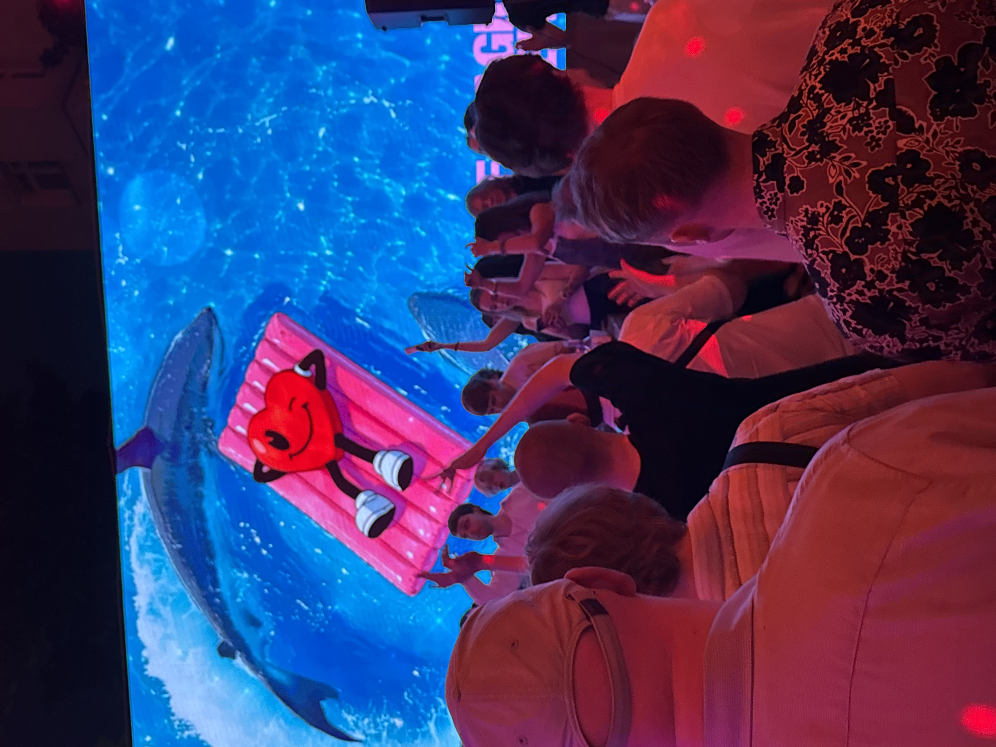 A group of people is gathered around a large screen displaying an animated image. The animation depicts a cartoon heart-shaped character lounging on a pink raft in blue water with a dolphin swimming beneath. The setting is indoors, possibly a public space in Moscow, Russia, although the specifics are not deducible from the scene. The people appear to be engaged with the screen's content.