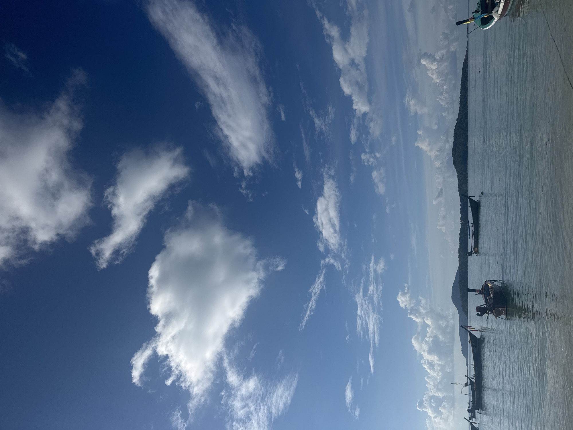 The image depicts a tranquil seascape in Rawai, Thailand. A clear blue sky, adorned with scattered white clouds, stretches across the frame. The calm, shallow water reflects the sky, creating a serene atmosphere. Several longtail boats, characteristic of Thailand, are moored in the water, their wooden hulls and distinctive prows visible. In the background, a verdant island with rolling hills rises from the horizon. The foreground shows the sandy shore of Rawai beach. The scene is devoid of people, suggesting a peaceful and unpopulated environment.