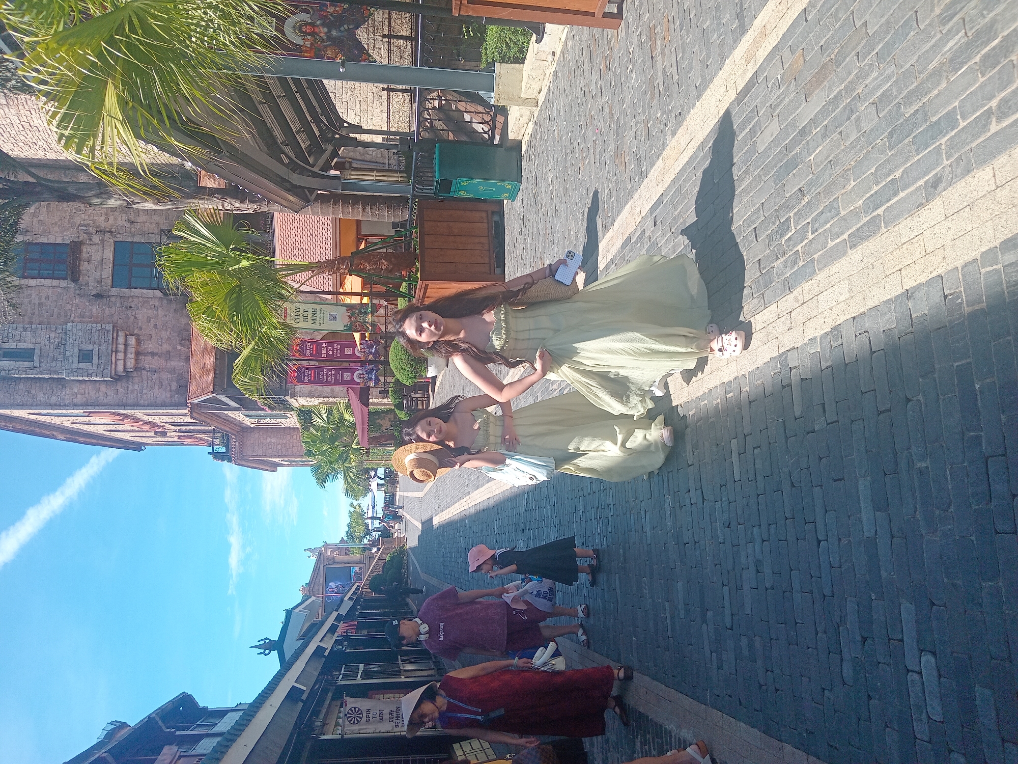 The image captures a cobblestone street scene in a tourist area, likely within the city of Da Nang, Vietnam, given the architectural style and tropical vegetation. The sun is shining brightly, casting distinct shadows on the pavement. On the right side of the frame, two young women are walking away from the camera, both wearing flowing, pale green dresses. The woman in the foreground is carrying a straw bag and a smartphone. The woman behind her is holding a straw hat. To the left, an adult woman wearing a dark red dress and a conical hat is walking towards the camera, accompanied by a man in a purple t-shirt and shorts, who is wearing headphones around his neck and carrying shopping bags. A young child in a dark dress and pink hat is walking between them. The buildings on either side of the street feature stone facades and are adorned with tropical plants, including palm trees. Banners with Asian text are visible on some of the buildings. A green trash receptacle is positioned near the building on the right.