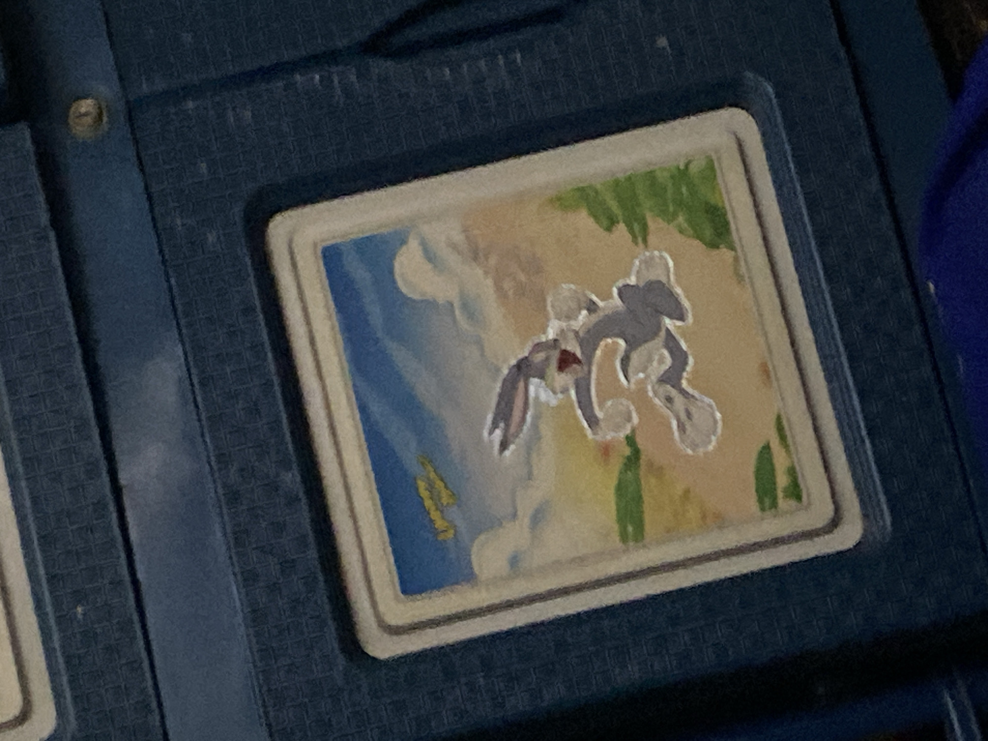 A close-up shot of a blue textured surface shows a framed image of Bugs Bunny in mid-air, seemingly running or jumping. The cartoon character is gray with white gloves and feet, and red inside its ears. The background depicts a sky with white clouds and a yellowish airplane. Green brushstrokes suggest foliage in the foreground and to the right of the image. The frame around the image is white and appears to be slightly raised from the blue surface. The blue surface itself has a grid-like texture. There is no visible indication of the city of Ta', Indonesia.