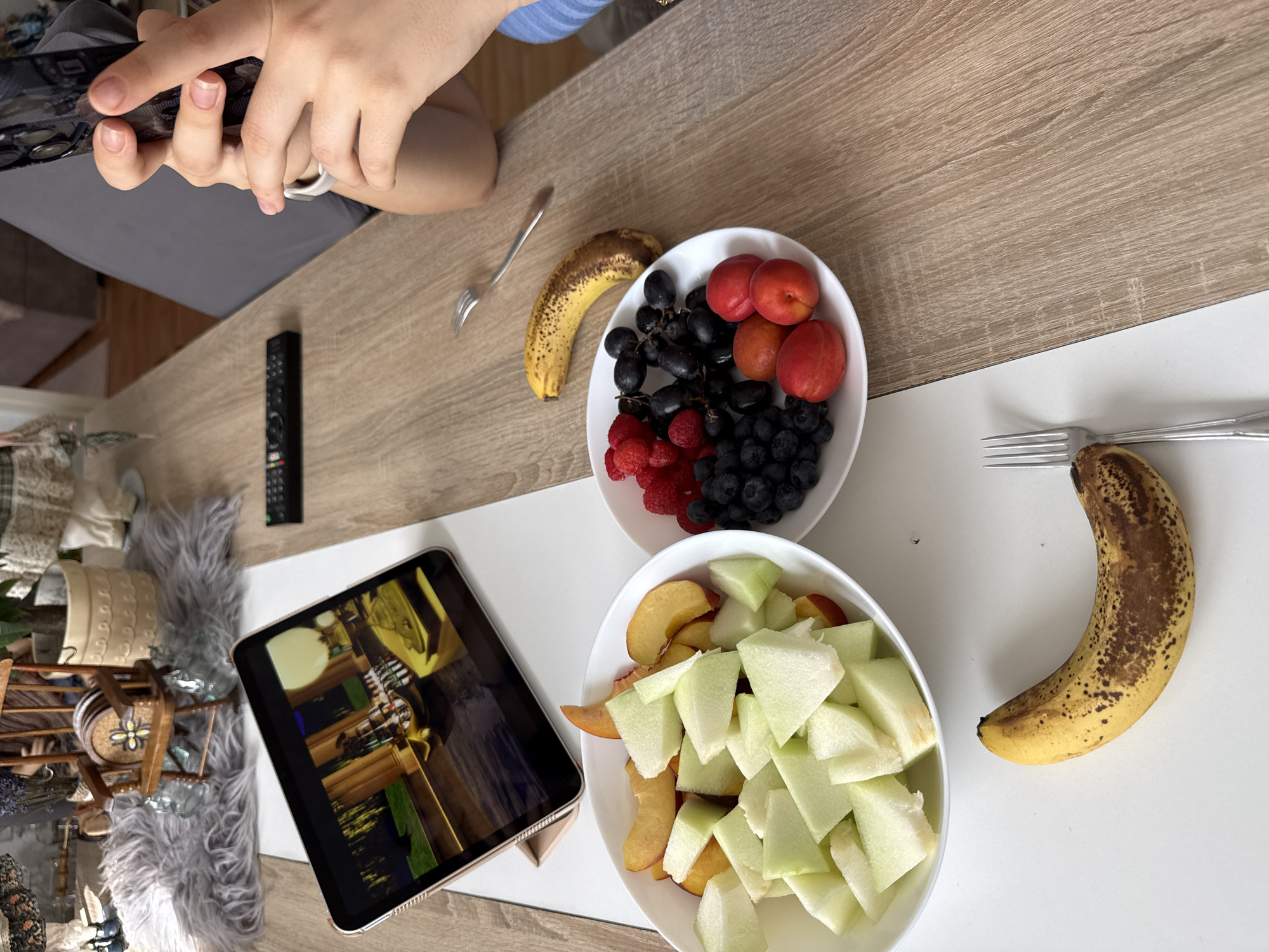 The image depicts a dining table at an indoor setting, possibly a residence. On the table, there are two white bowls filled with various fruits, including chopped melon and peaches in one, and raspberries, blueberries, grapes, and apricots in the other. A whole banana lies on the table beside one of the bowls. An iPad is propped up on a stand, displaying an image. A remote control is also visible on the table. In the foreground, a person's hands are holding a smartphone, suggesting they are taking a picture or recording a video of the scene. In the background, a small wooden rocking chair with a fluffy rug in front of it can be seen, along with some decorative items. The overall impression is a casual setting where someone is enjoying a fruit platter while possibly watching content on their tablet or capturing the moment with their phone. The location in the city of Hanau, Germany, is not indicated by any visual elements in the image.