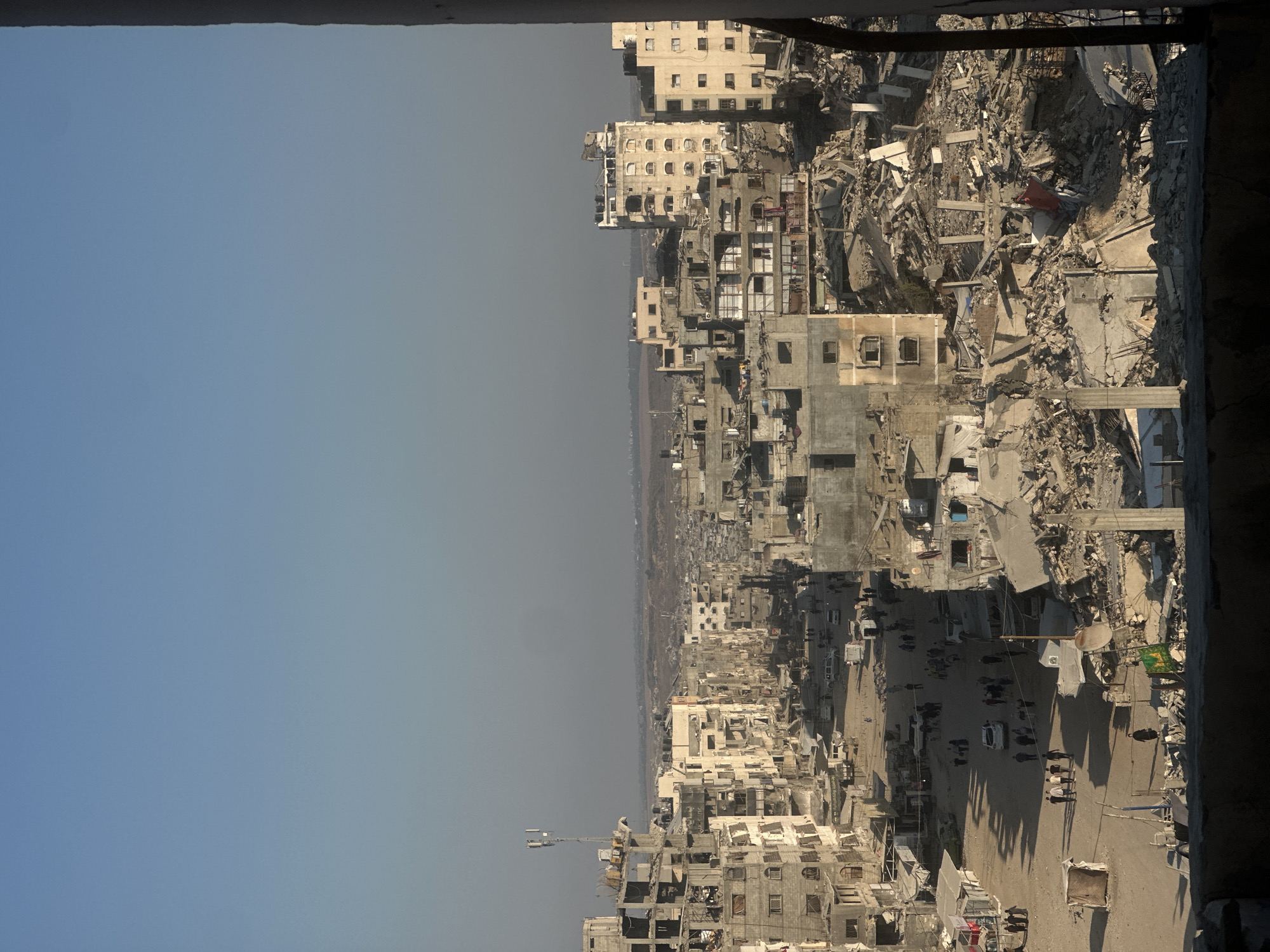An elevated, eye-level, medium shot captures a street scene in the city of Gaza, Palestinian Territories, with numerous buildings in varying states of destruction. The sky above is clear and a pale blue. In the foreground, a dark, shadowy balcony railing frames the bottom portion of the image. The street is a dusty, light brown, with a mix of intact and severely damaged buildings lining both sides. Some buildings show significant structural damage, with collapsed walls, exposed rebar, and missing sections. Others appear partially intact, with windows and balconies visible. Rubble and debris are scattered throughout the scene, particularly on the right side of the image, where entire structures have been reduced to piles of concrete and twisted metal.
A few vehicles, including white vans and cars, are parked or moving along the street. Several groups of people are walking and gathered on the street and sidewalks. Some individuals appear to be inspecting the damage or navigating the debris. The overall impression is one of devastation and ongoing activity amidst widespread destruction. The lighting suggests it is daytime, with shadows cast by the buildings indicating the sun is positioned to the left. The distance reveals a dense urban landscape stretching to the horizon.