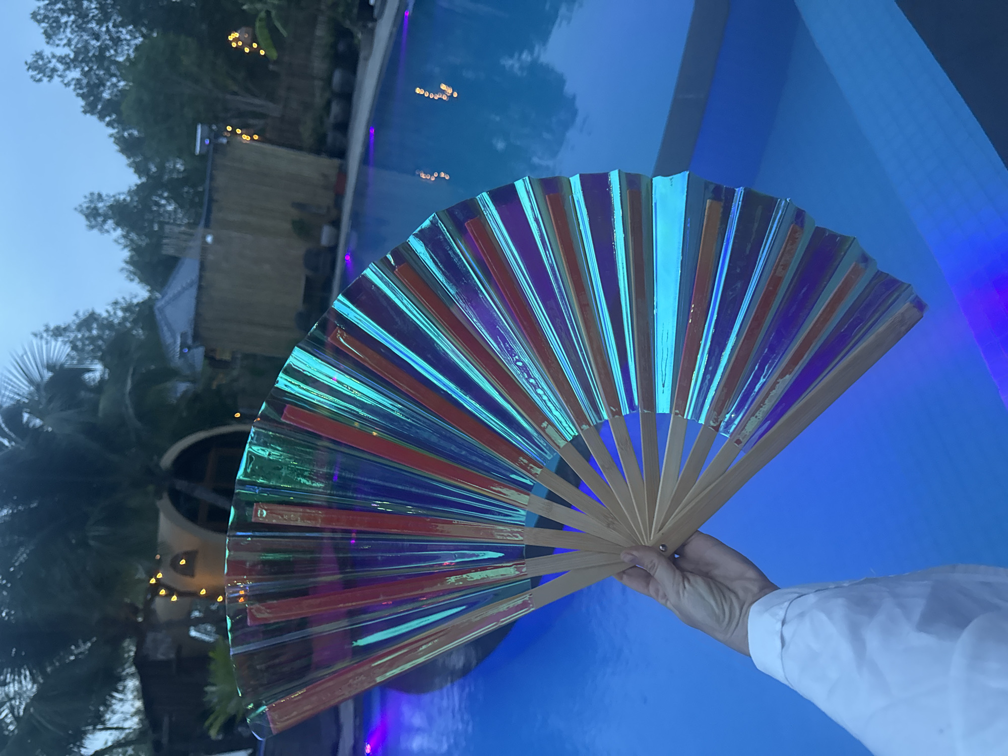In Mũi Né, Vietnam, a person holds a semi-circular, iridescent fan made of clear plastic with red and blue hues. The fan is open, revealing wooden sticks along its base. The fan is held in front of a swimming pool illuminated with blue and purple lights. In the background, palm trees and rustic structures are visible, suggesting a tropical resort setting. The sky is overcast, indicating it is likely late evening or early morning.