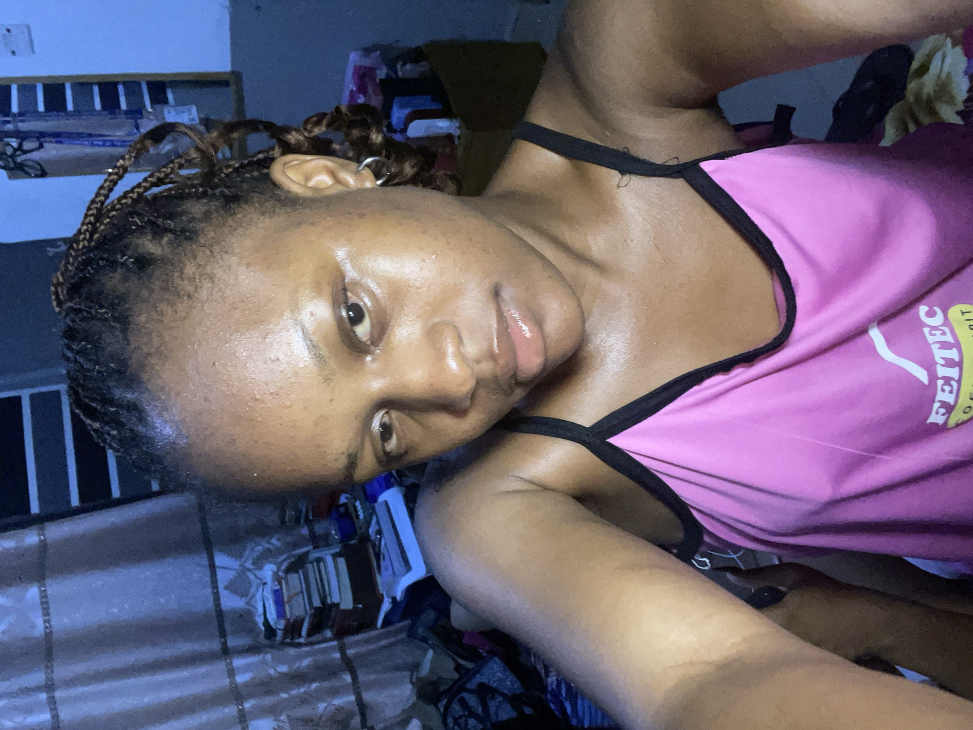 A medium shot captures a young woman with braided hair and a pink tank top, posing for a selfie in what appears to be a bedroom. She has a direct gaze and her skin is slightly oily. The background is cluttered with stacked books, fabric, and a mirror reflecting a window with horizontal blinds. The overall lighting suggests an indoor setting, possibly at night. The location is identified as Rafin Par, Nigeria.