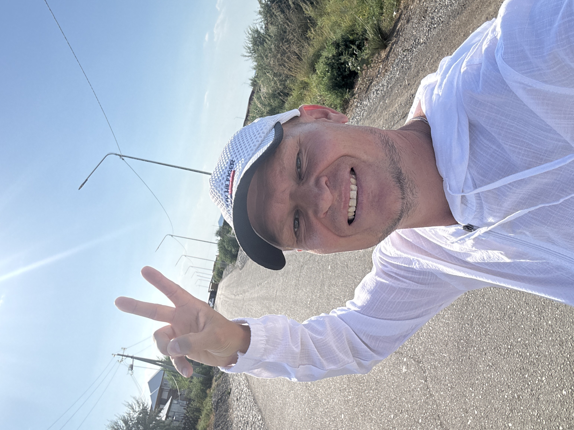 In a selfie taken on a sunny day, a man with a cap and white hoodie is standing on a paved road in Zharbulak, Kazakhstan. He is smiling and making a peace sign with his right hand. The road appears to be in a rural or suburban area, with houses and vegetation visible in the background. Utility poles and streetlights are also present along the road. The man is the primary subject of the image.