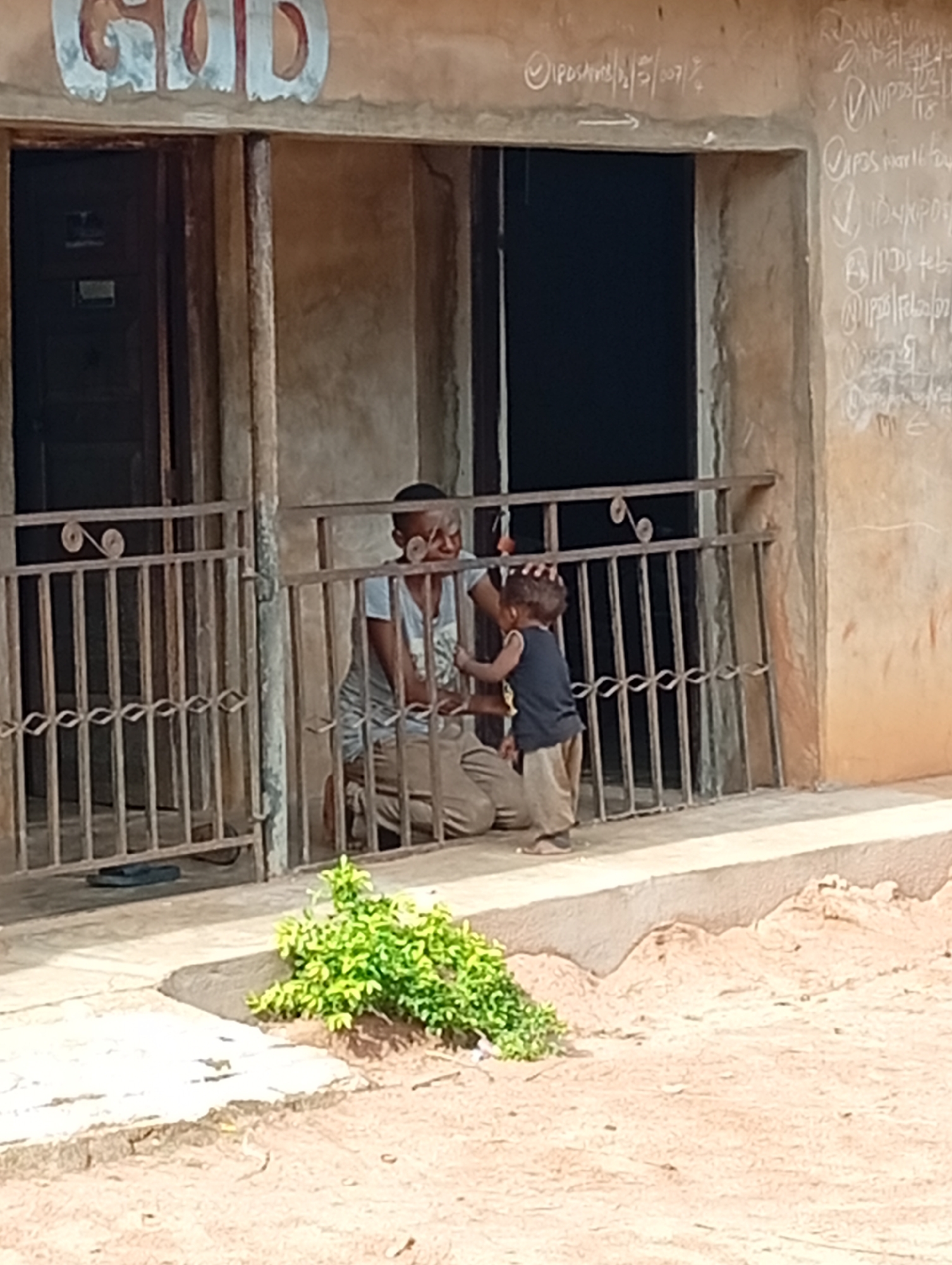 In Adda, Nigeria, a man kneels behind a metal fence, facing away from the camera, while a young child stands facing him. The man is wearing a light-colored t-shirt and khaki pants, and has his hand placed on the child's head. The child, wearing a dark sleeveless shirt and light-colored pants, is looking up at the man. The scene takes place on the ground level of a building with a weathered facade. Above the entrance to the building, the word "GOD" is painted in large, white, stylized letters with red and blue accents. The wall to the right of the entrance is covered in chalk writing, some of which appears to be names and dates. A small patch of green foliage grows near the fence in the foreground. A pair of dark sandals lies on the ground behind the fence.