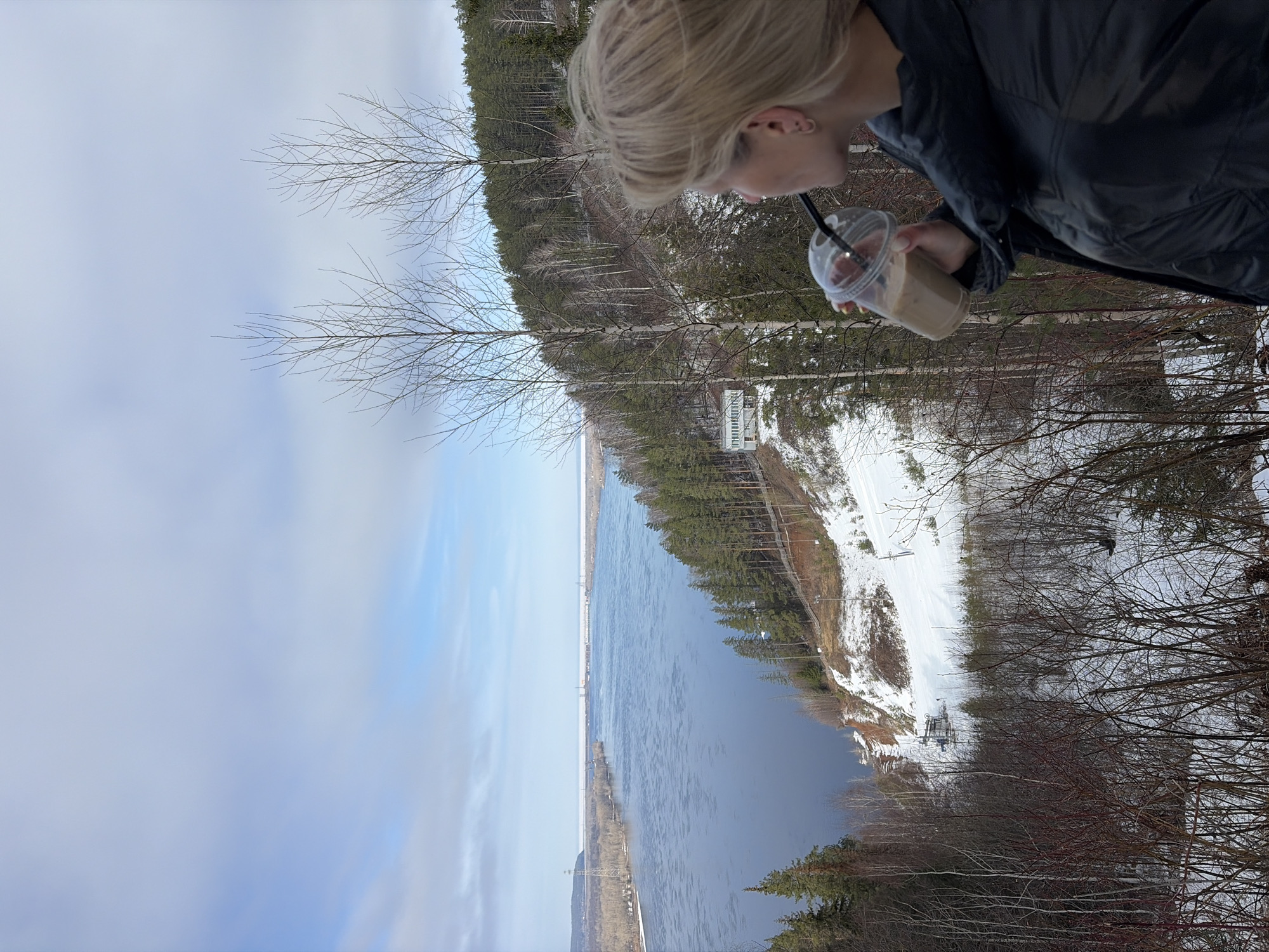 A woman with blonde hair and wearing a black jacket is drinking from a clear plastic cup with a black straw. The drink appears to be a latte or similar coffee beverage. She is standing outdoors, overlooking a wide river.
Across the river is a landscape with snow-covered areas and dense forestation, likely during late winter or early spring. A ski lift or similar infrastructure is visible on the opposite bank. The sky is overcast, with a mix of blue and grey clouds.
The location appears to be a vantage point overlooking the Volga River near Noviy, Russia. Specifically, the woman seems to be standing near a ski area on the right bank of the river, looking across towards potential residential or recreational areas.