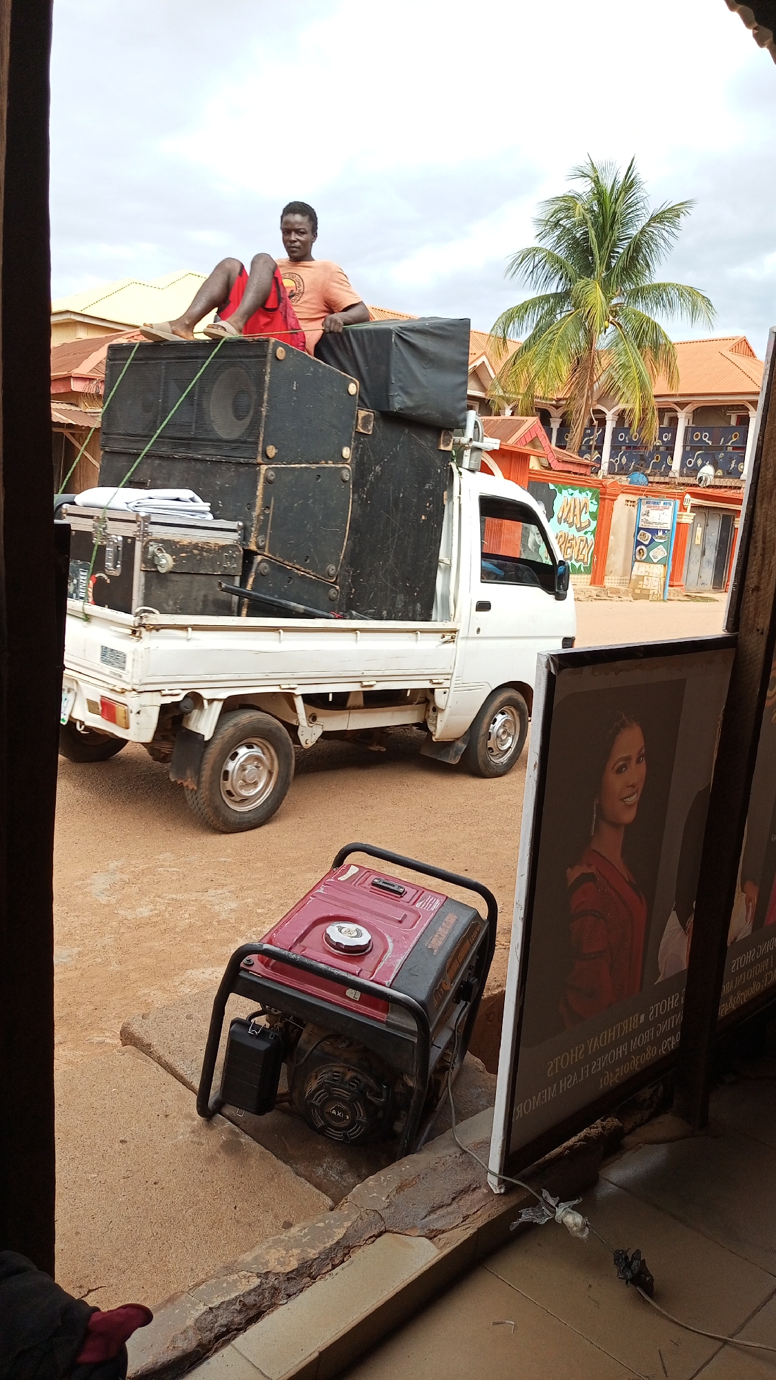 A man sits atop a stack of large black speakers loaded onto the back of a small white truck. The man is wearing an orange shirt, red shorts, and sandals. The truck is parked on a dirt road in Sabon Tasha, Nigeria. Behind the truck is a building with an orange roof and decorative trim. In the foreground, near the bottom of the image, there is a red and black generator. Part of the scene is obscured by a signpost in the foreground.