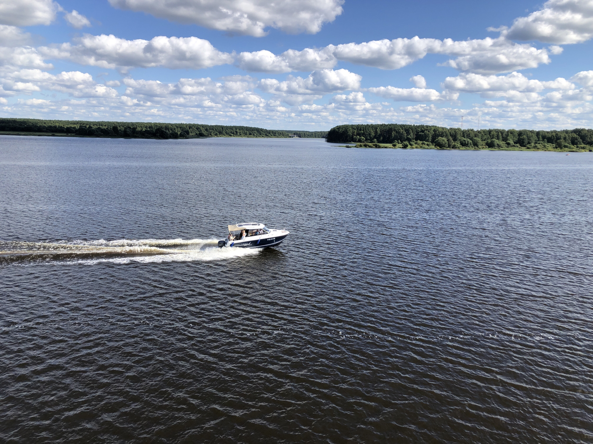 A motorboat travels across a wide, dark blue river under a bright blue sky filled with fluffy white clouds. The boat, which is white with blue trim, has a canopy and several people on board. It is creating a white wake as it moves from left to right across the frame. The riverbanks are lined with dense green trees. The image appears to be taken from an elevated position, looking down at the boat and the river. The scene is serene and picturesque, suggesting a pleasant day for boating. The location is the Volga River near the city of Myshkin, Russia.
