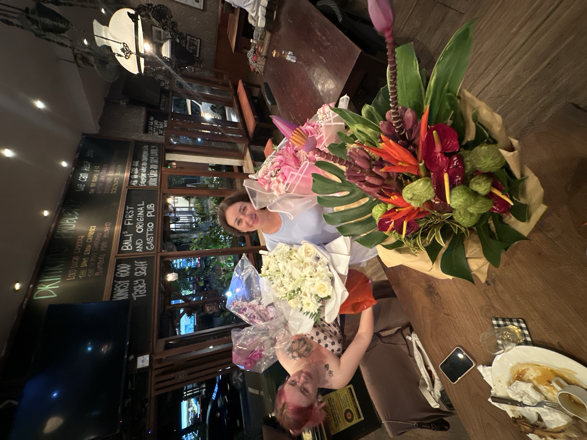The image depicts the interior of a gastro pub called "Bali's First and Original Gastro Pub", possibly located in North Kuta, Indonesia, as suggested by the writing on the chalkboard. Two women are seated at a table. One woman, with pink hair, a sleeveless top, and visible tattoos, is holding a bouquet of white roses. The other woman, wearing a light blue shirt, is holding a bouquet of pink flowers. A large floral arrangement with tropical flowers and greenery is on the table. A partially eaten meal, silverware, a cellphone, and a checkered napkin are on the right of the table. In the background, there is a TV, a chalkboard menu with a list of drinks, and the pub's name. Windows are visible behind the people. The pub is dimly lit.