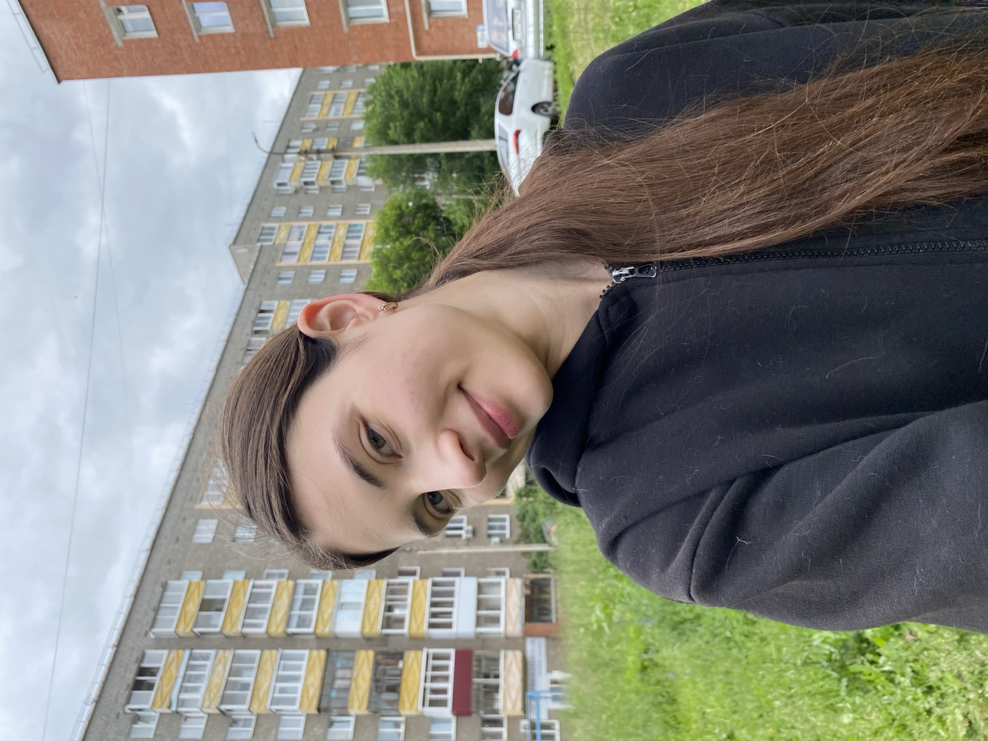 A young woman with long, brown hair pulled back into a ponytail takes a selfie outdoors. She is wearing a black hooded sweatshirt. Behind her, a grassy area is visible, with several apartment buildings and a few cars in the background. The architecture of the apartment buildings suggests a Soviet-era design, commonly found in Russian cities. The sky is overcast with clouds. The overall setting is a residential area in the city of Votkinsk, Russia.