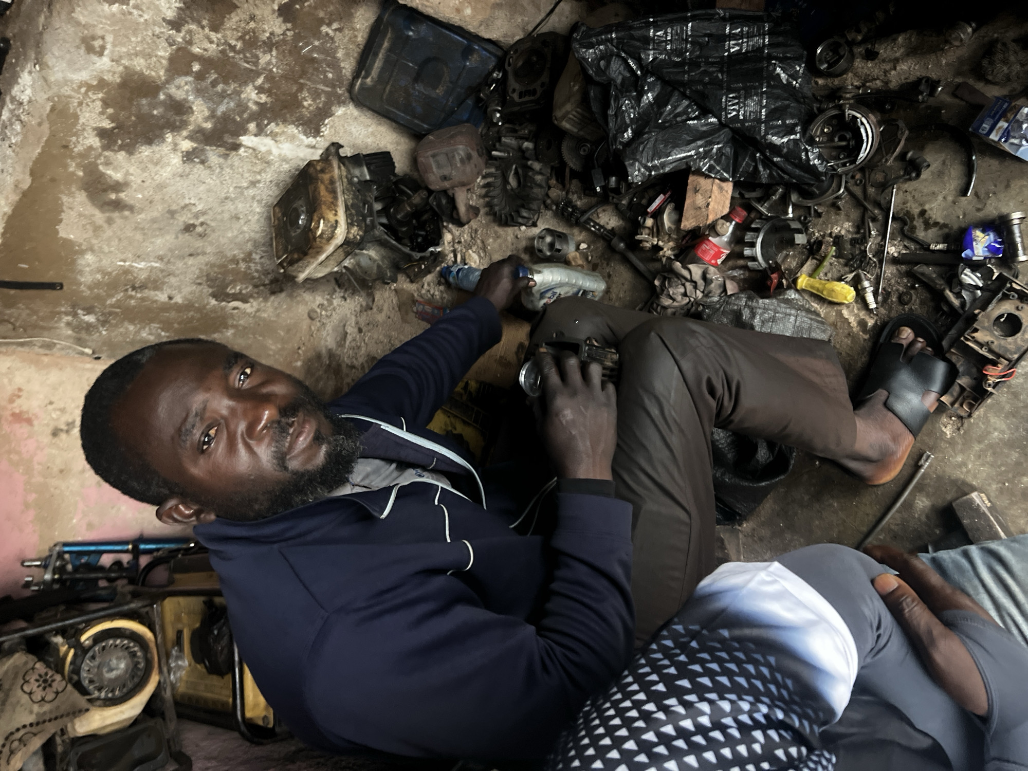 A Black man is kneeling in a cluttered workshop, surrounded by engine parts and tools. He is wearing a dark blue jacket and brown pants, and has a beard. He is holding a can in his left hand and looking at the camera. The workshop appears to be dimly lit and has concrete walls. To his left, another person is partially visible, wearing a black and white patterned garment. The background contains a generator and various mechanical components, suggesting an automotive or engine repair environment. The scene is located in the city of Jos, Nigeria.
