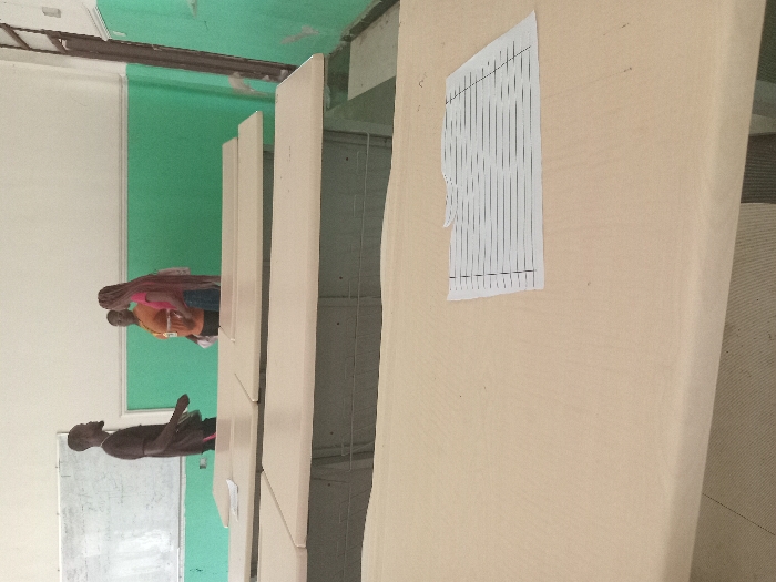 The image depicts a classroom setting, potentially in Umueshi, Nigeria. Several light-colored desks are visible, with a printed sheet of paper attached to one. People are present, some appearing to be seated or standing around the desks, suggesting a learning environment. A whiteboard is also visible, further supporting the classroom context. The overall scene indicates a space dedicated to education, likely within a school setting.