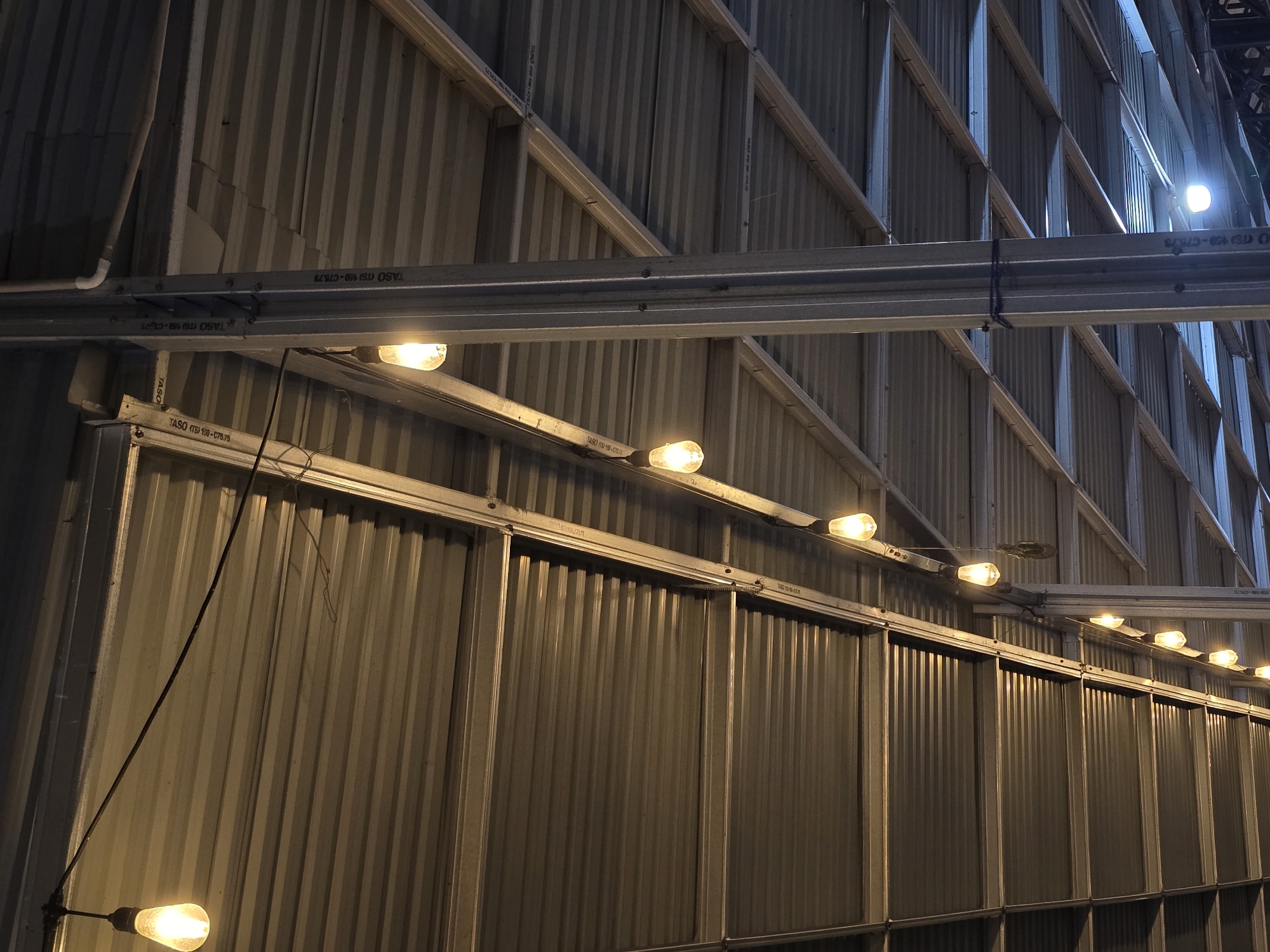 The image shows a view looking up at the corrugated metal ceiling of an outdoor structure, possibly a market or covered seating area, illuminated by a string of incandescent Edison bulbs. A large vertical metal support column is visible on the right side of the frame. The ceiling is supported by a series of metal beams and trusses. The lighting creates a warm, ambient glow. The scene appears to be located in Dayah Kruet, Indonesia.