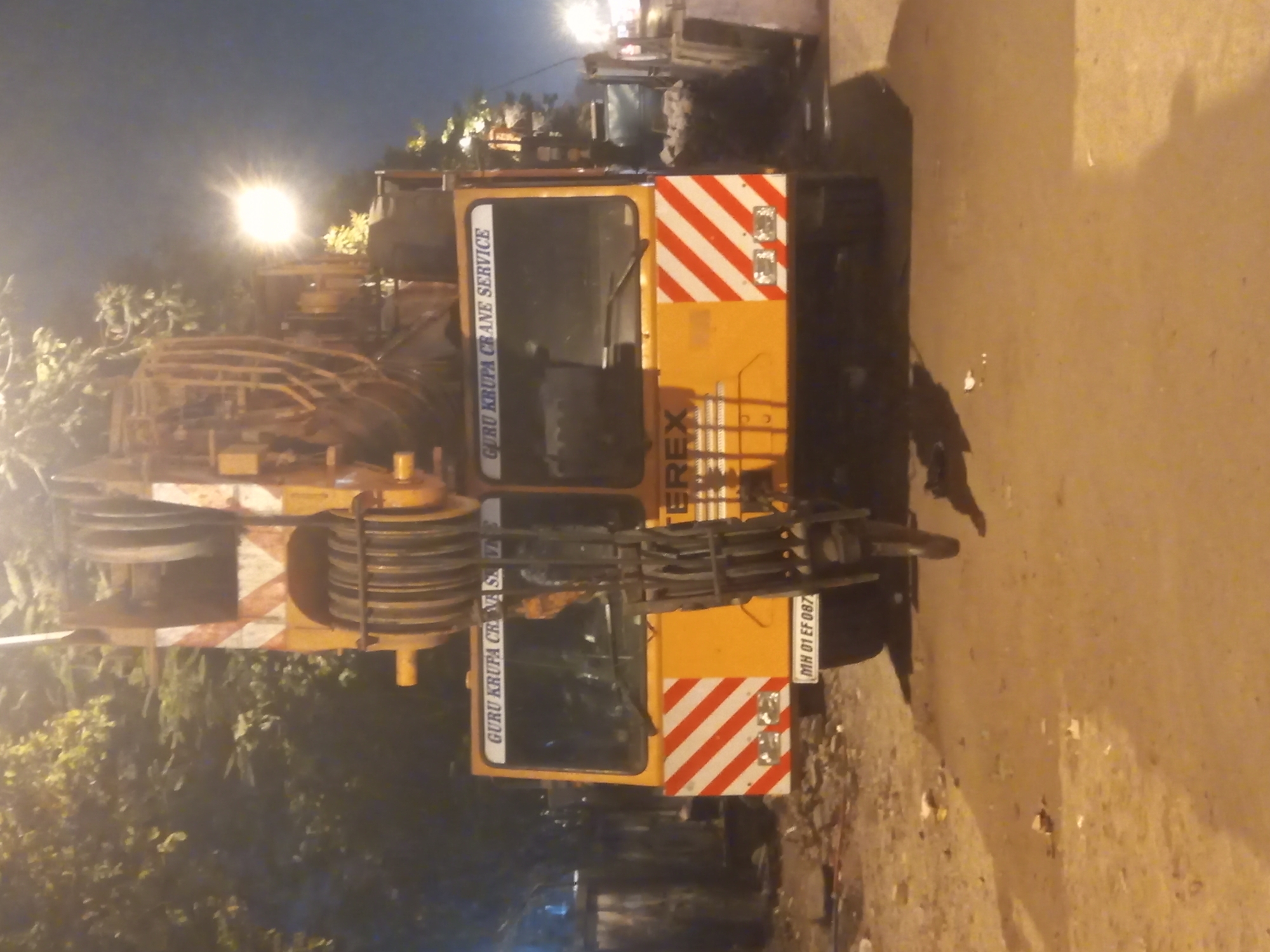 The original image, which is rotated 90 degrees clockwise, shows a large, yellow mobile crane parked on a street at night. The sky is dark, and the scene is illuminated by artificial light sources, including a prominent, bright street lamp in the background on the left.
The main subject is the yellow mobile crane, which has a substantial boom structure and pulleys/blocks at its end. The crane's cab is visible, along with parts of its chassis and wheels. The rear of the crane features red and white diagonal striped hazard markings and a license plate. Trees or dense foliage are visible on the left side of the frame, in the background relative to the crane. The ground surface to the right of the crane appears to be a paved road or street.
Visible text on the crane includes "GURU KRUPA CRANE SERVICE" printed vertically on the side of the cab and again on the lower section. The brand "TEREX" is partially visible below the cab window. A license plate at the rear of the crane displays "MH 01 EF 887", indicating it is registered in Maharashtra, India.
The time of day is nighttime, evident from the dark surroundings and reliance on artificial lighting. There is no discernible activity taking place; the crane appears to be stationary or parked. No people are visible.