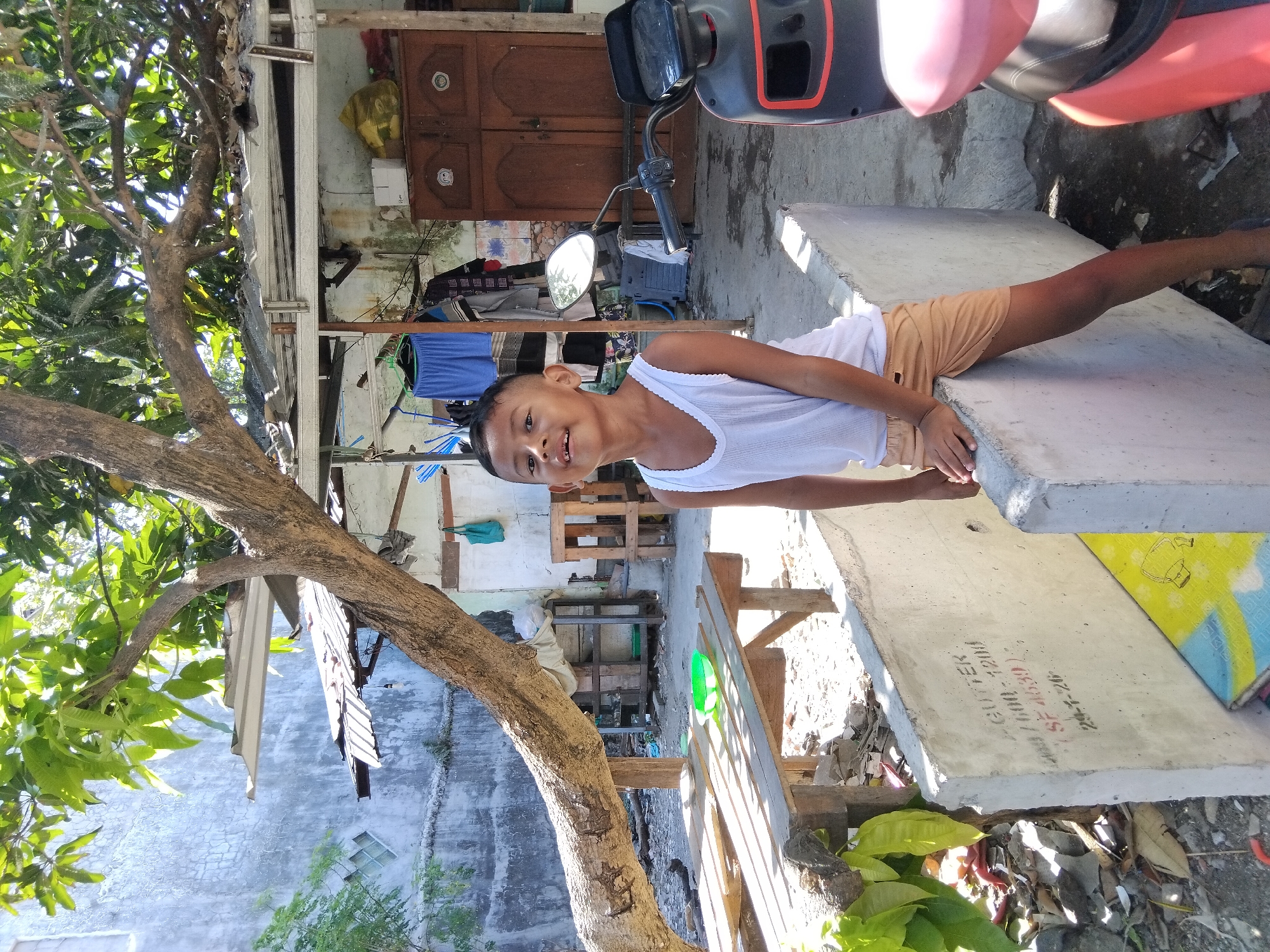 An outdoor medium shot captures a young boy smiling broadly as he leans on a concrete barrier in a rural Indonesian setting. He is wearing a white tank top and tan shorts. The background reveals a weathered wooden structure with a corrugated metal roof, reminiscent of typical rural architecture in Berbek, Indonesia. A tree with large leaves partially obscures the scene, and a motorcycle is parked to the right of the boy. The ground is a mix of dirt and scattered debris, with a wooden table and chairs in the foreground. The image appears to have been taken during daylight hours, with harsh sunlight casting distinct shadows.