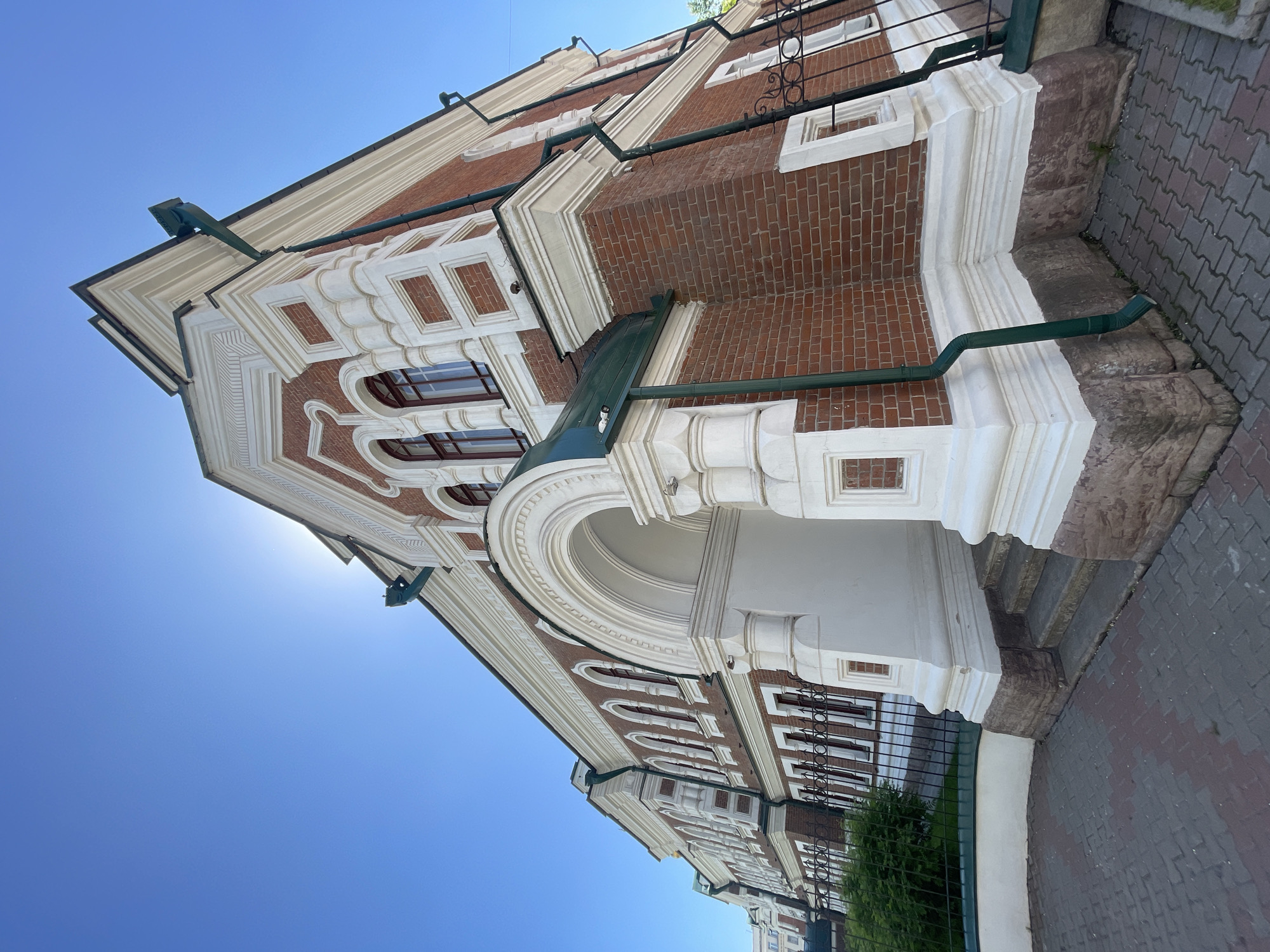 This media file captures a low-angle view of an ornate brick building facade under a clear, bright blue sky in Krasnoyarsk, Russia. The multi-story structure prominently features red brickwork complemented by extensive white architectural embellishments, suggesting a historical style such as Baroque or Neoclassical.
Notable details of the facade include elaborate stucco or plaster detailing around windows, cornices, and decorative pilasters. The windows are varied, with some featuring rounded arches and others rectangular frames, all with dark sashes. Several upper-story windows are adorned with decorative wrought-iron grilles. A prominent arched portico or entranceway is visible towards the center-right of the main image. Green metal gutters and downspouts are integrated into the building's exterior, contrasting with the red brick and white trim.
The time of day appears to be daytime, with a bright sun creating a glare in the upper left portion of the frame, indicating clear, sunny weather. The building stands on a visible stone foundation adjacent to a patterned grey paved sidewalk. A dark metal fence and some green foliage are visible at the lower right. No people or specific activities are depicted, allowing the architectural grandeur of the building to be the sole focus. There is no visible text.